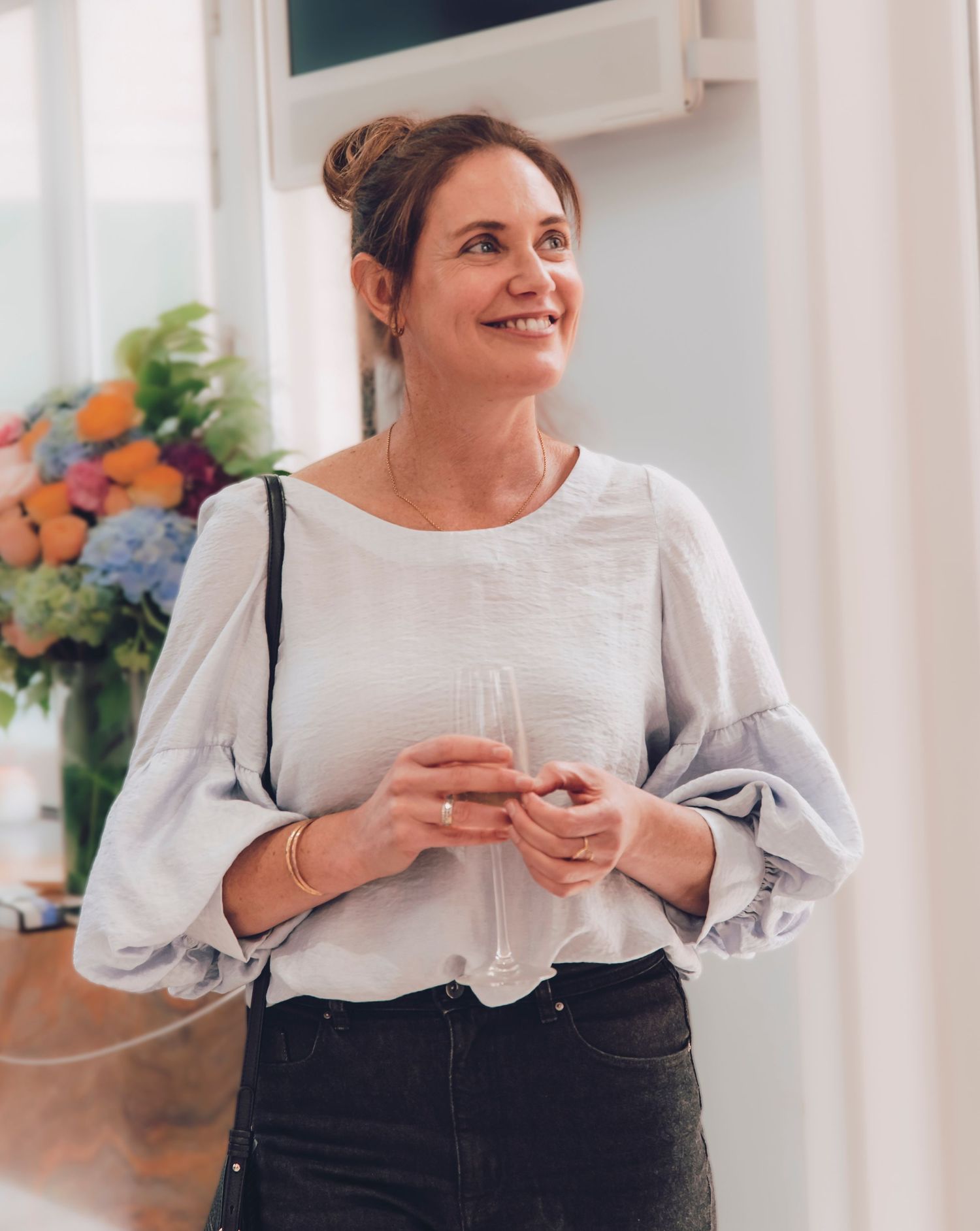 Woman in white blouse and black dress smiles while holding a coffee cup in a bright kitchen setting.