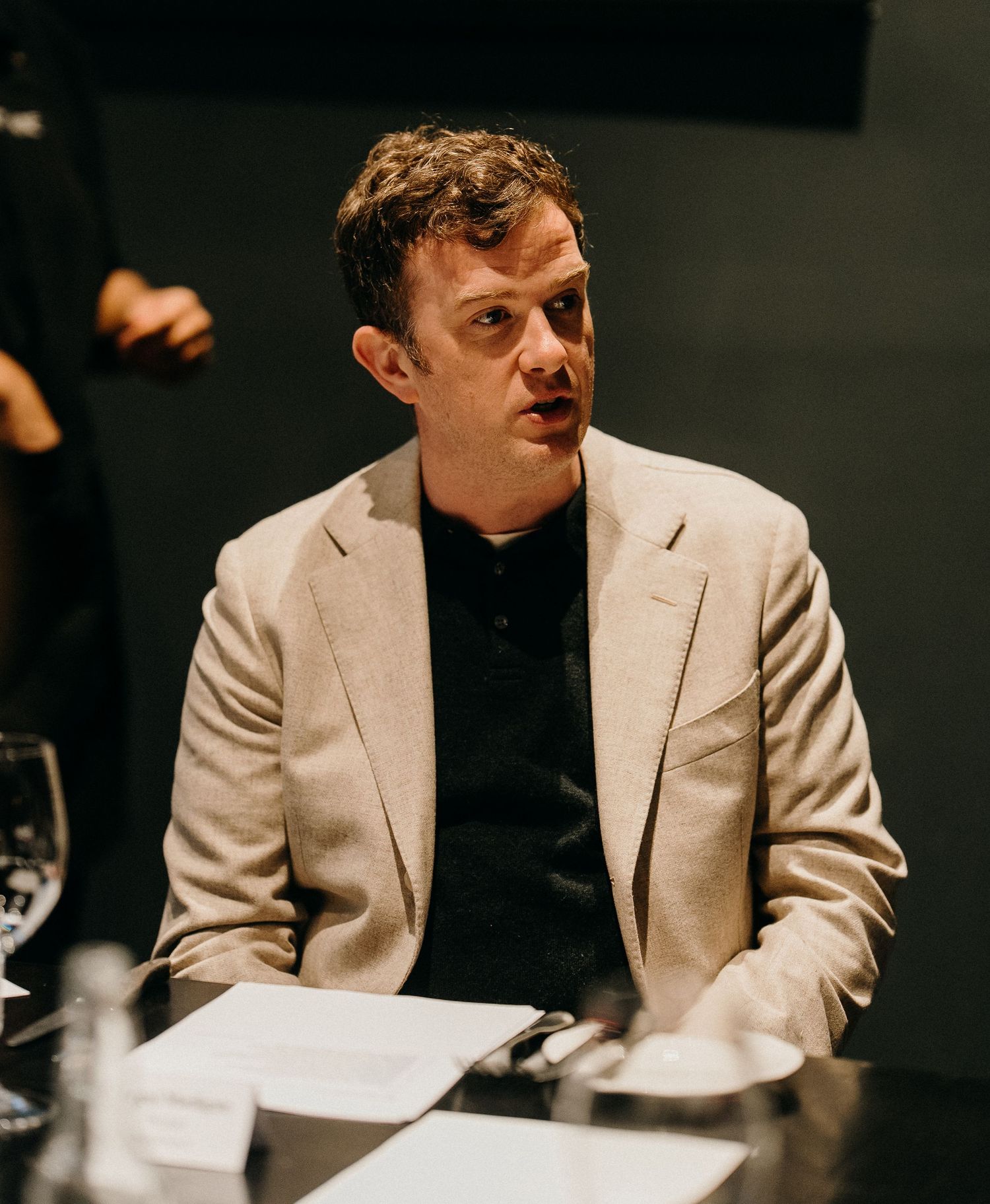 A person in a white blazer and black shirt sits at a table in a dark room during what appears to be a meeting.