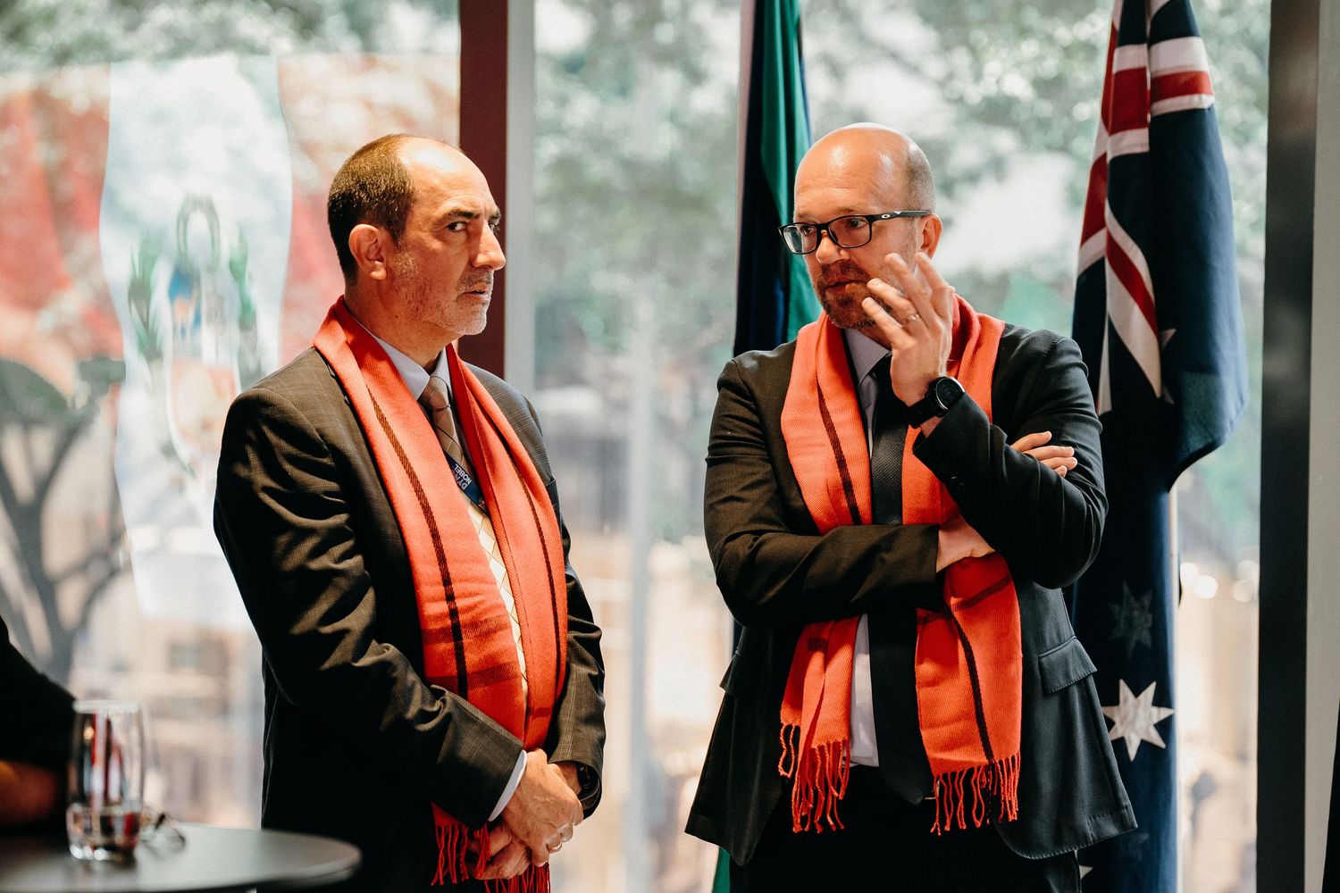 A row of people in black suits and orange scarves stand near large windows during an indoor event.