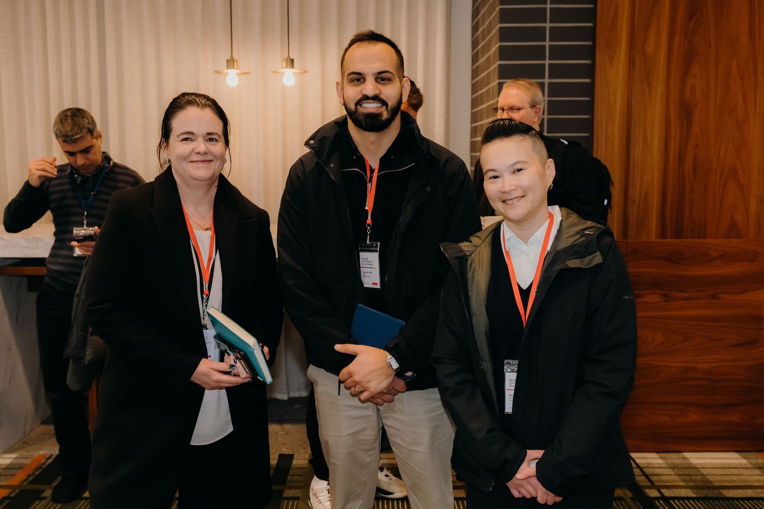 Three people wearing conference badges stand together in a wood-paneled room with pendant lighting.
