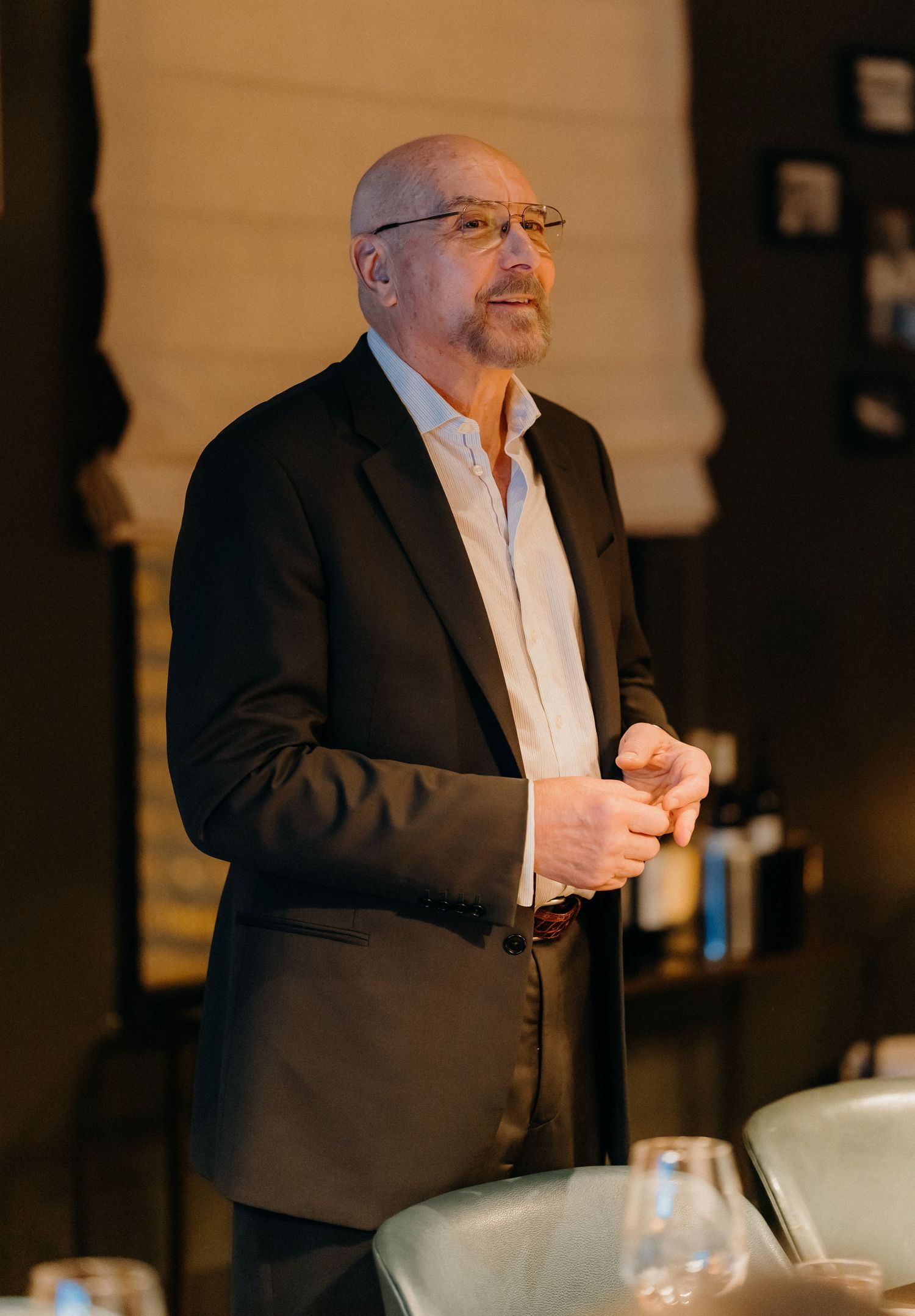 A person in a black blazer and white shirt stands thoughtfully in a dimly lit room with wine glasses visible.