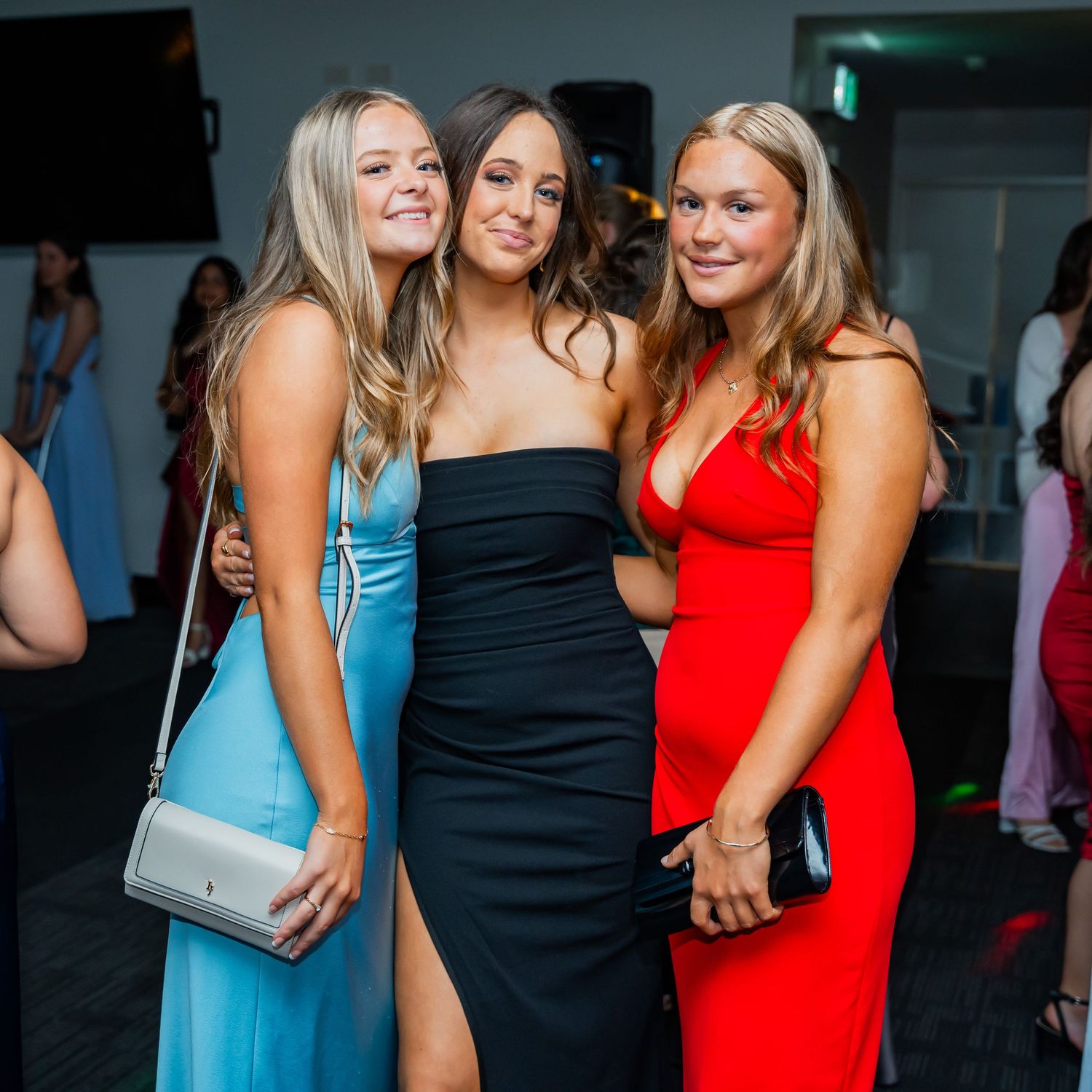 Three friends wearing formal dresses in light blue, black and red pose together at an evening event.