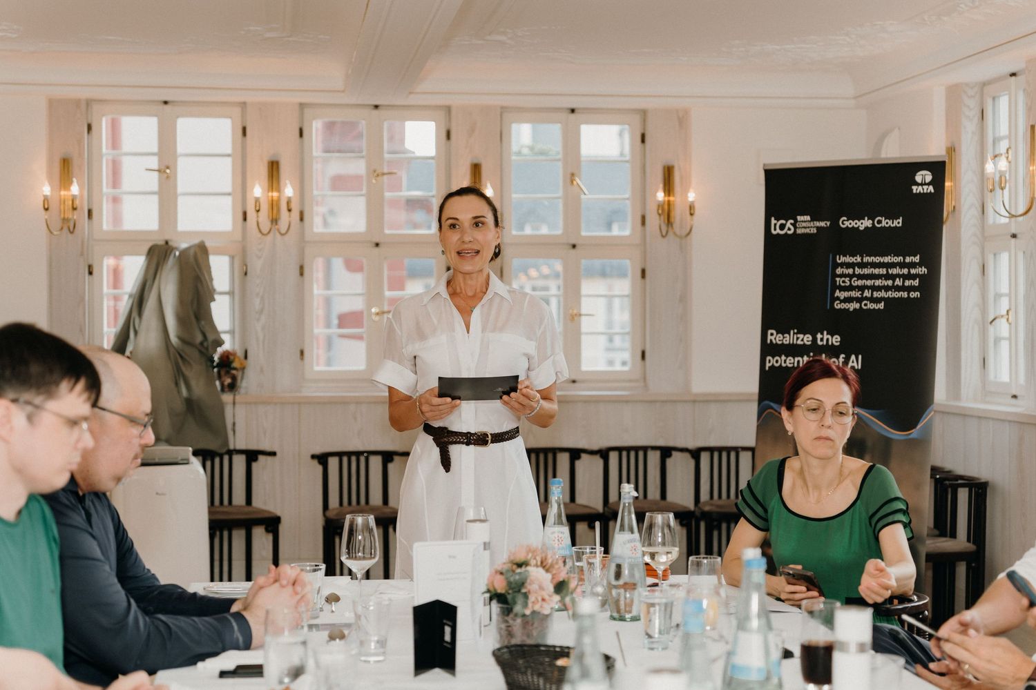 Woman in white dress presenting at a Google Cloud business event to seated attendees in an elegant room.