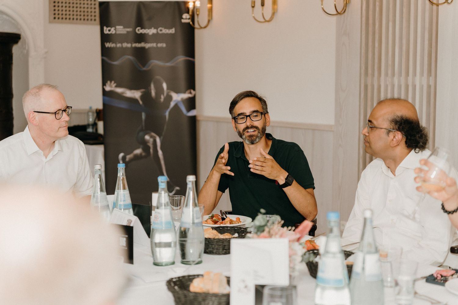 Three men engaged in lively discussion at a business dinner table with Google Cloud branding banner in background.