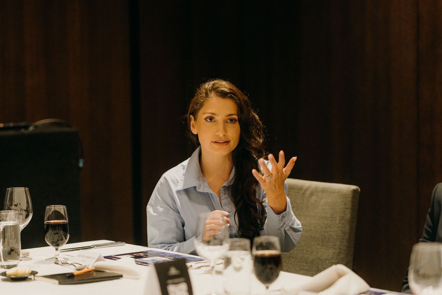 Woman gesturing while speaking at a formal dinner table with wine glasses in an elegant restaurant setting.