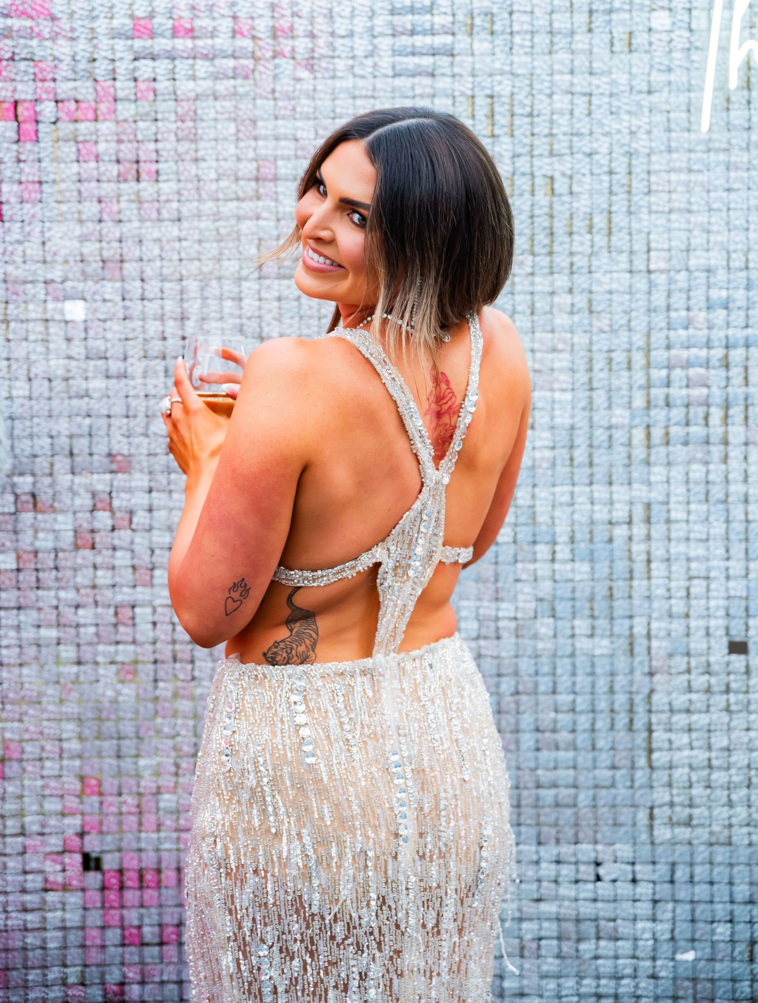 A woman in a sparkly white fringe dress poses and dances against a tiled wall backdrop.