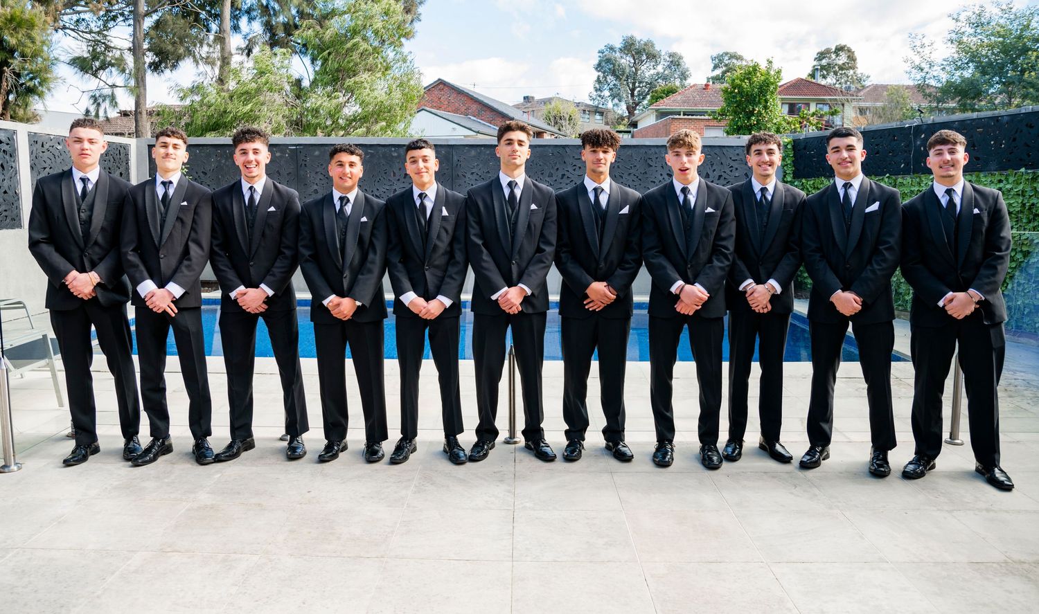 Wedding party in black suits stands in a long line outdoors with greenery in the background.