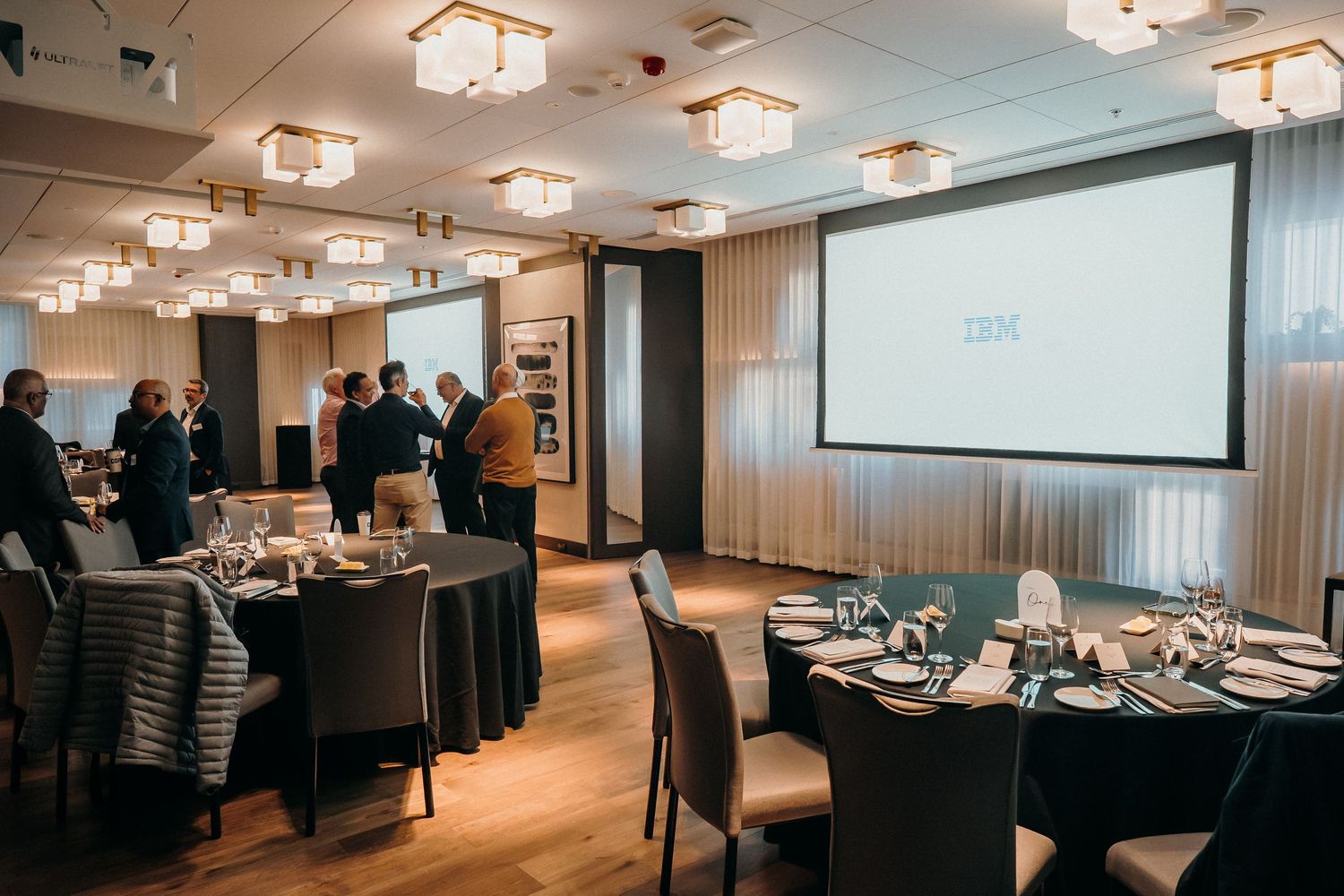 A modern conference room with round dining tables and elegant place settings illuminated by contemporary ceiling lights.