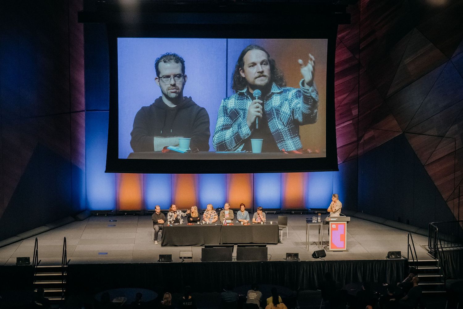 Two speakers present on stage at a technology conference with large projection screens displaying their content.