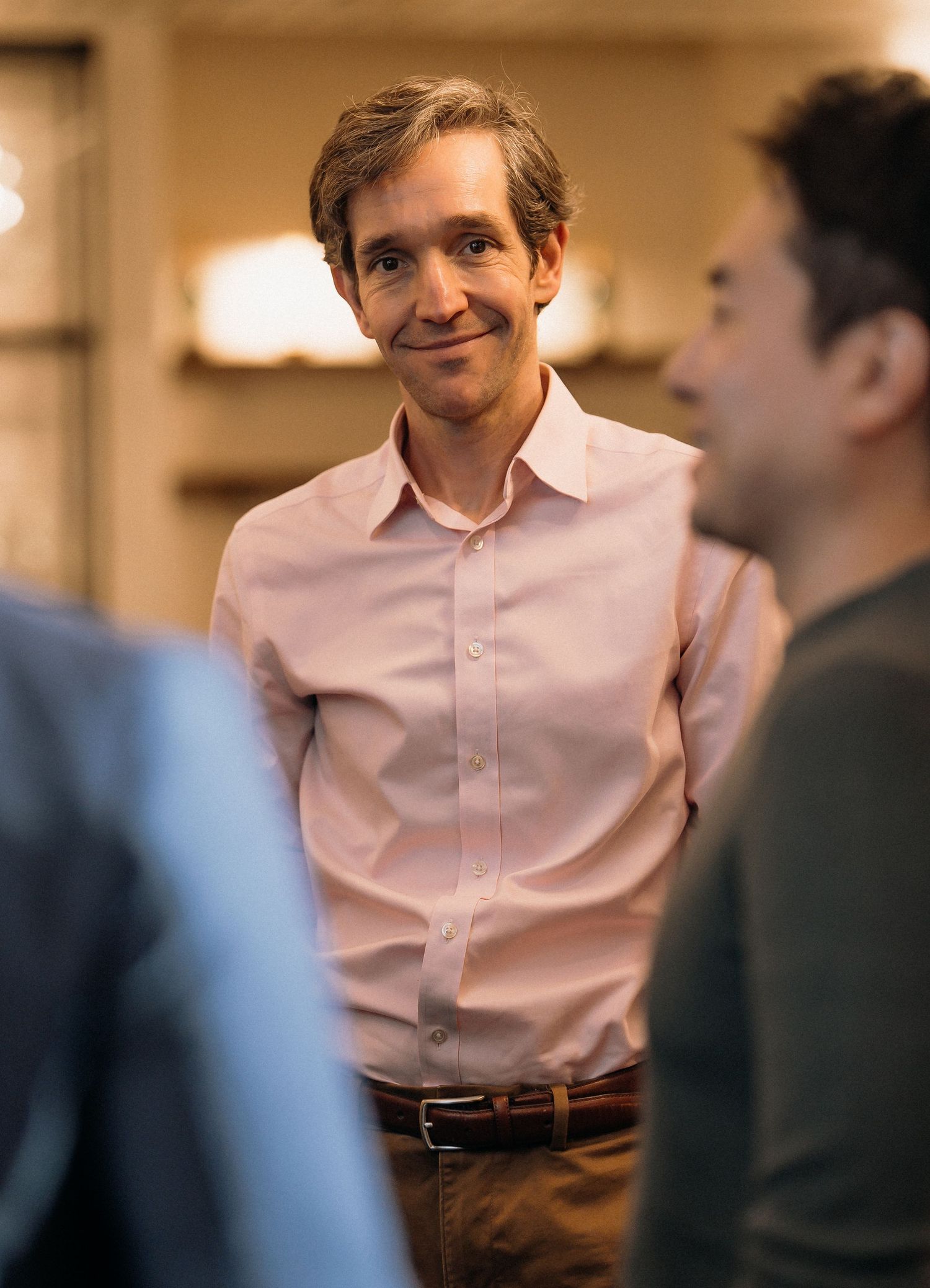 Person in pink dress shirt engaged in animated conversation with others in warm-lit indoor setting.