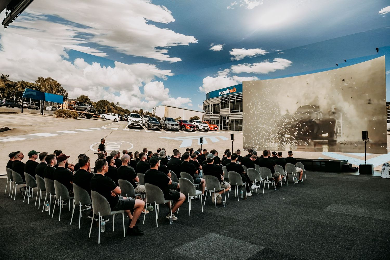 Outdoor audience seated in white chairs watches presentation on large wall screen under bright blue sky with puffy clouds.