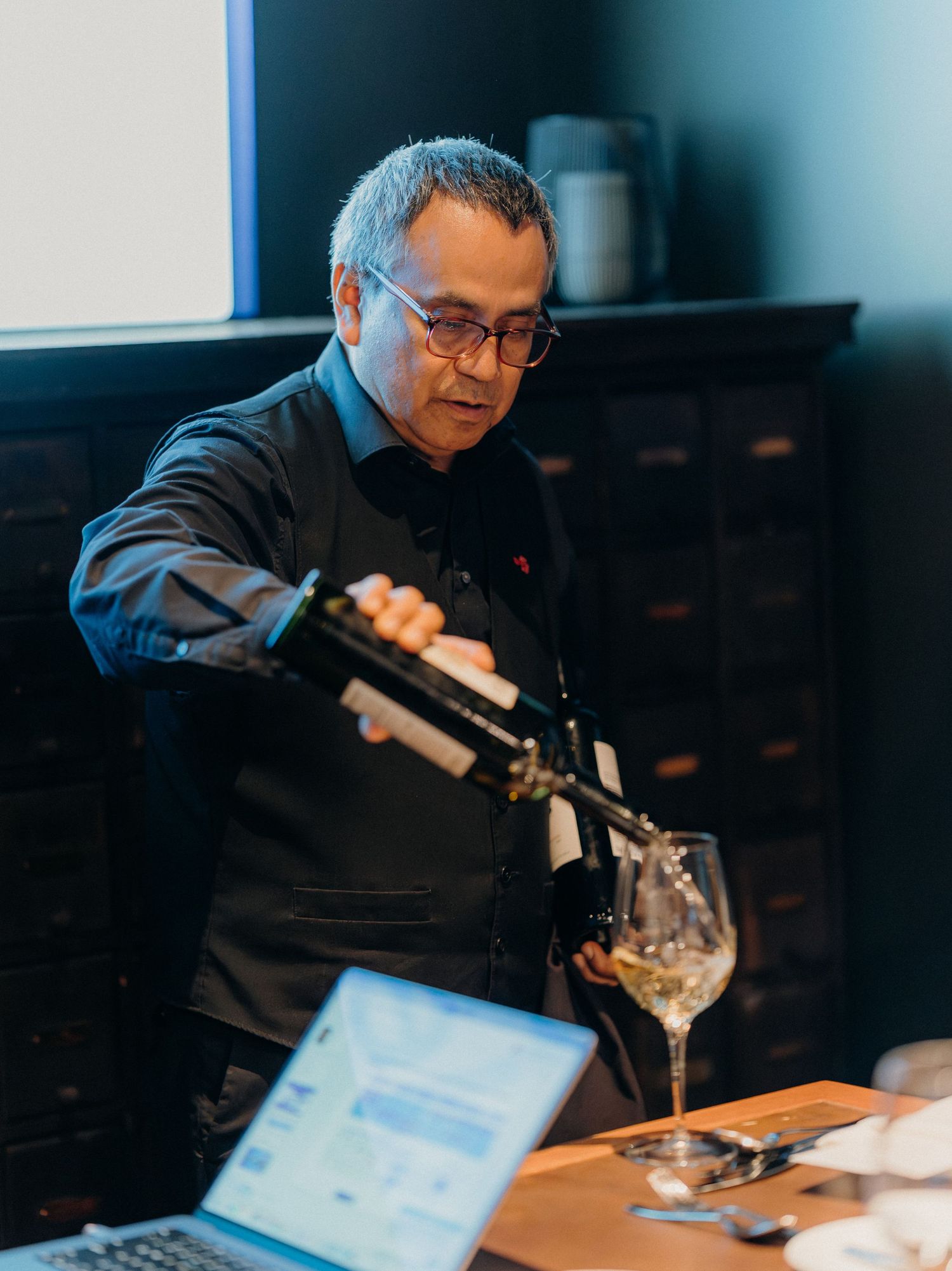 Professional sommelier pouring white wine into a glass at a tasting event in a dimly lit setting.
