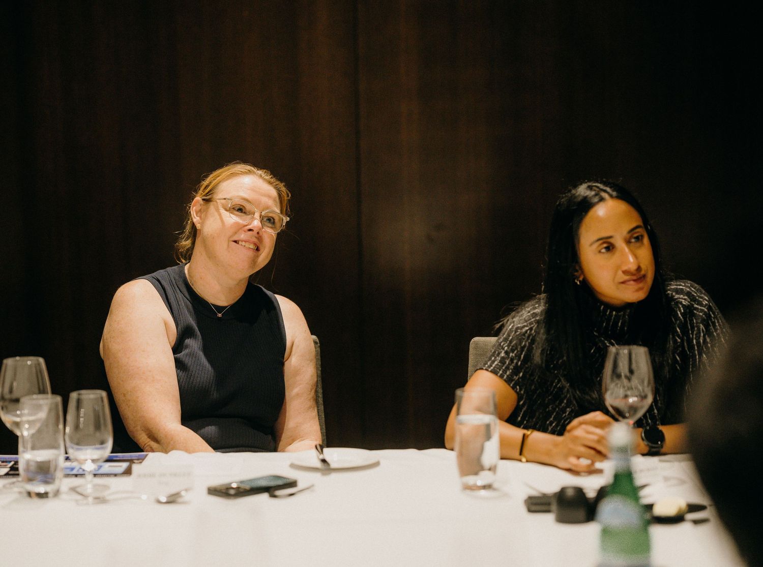 Two women sitting at a formal dining table with wine glasses, engaged in conversation at an indoor event.
