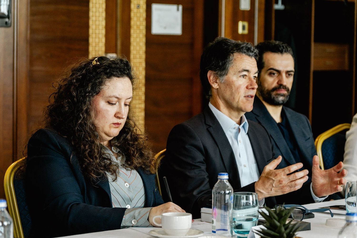 Three professionals seated at a conference table engaged in discussion, with a man gesturing while speaking at a formal meeting.