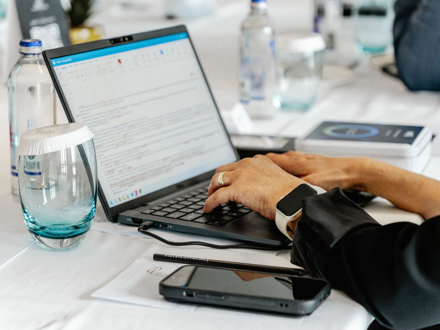 Person typing on laptop at conference table with water bottles, smartphone, and notepad nearby.