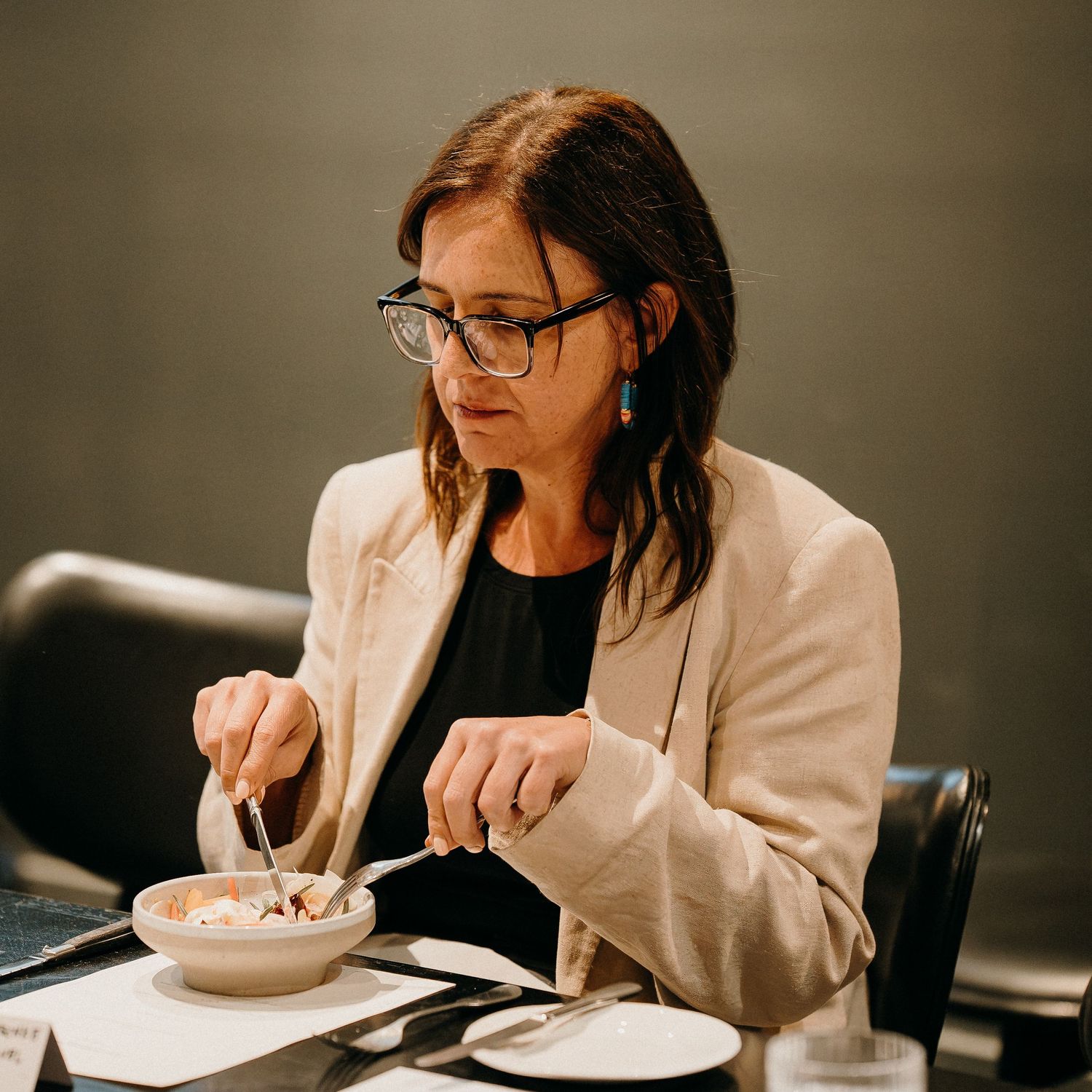 Person in white blazer eating soup from white bowl at dimly lit restaurant table.