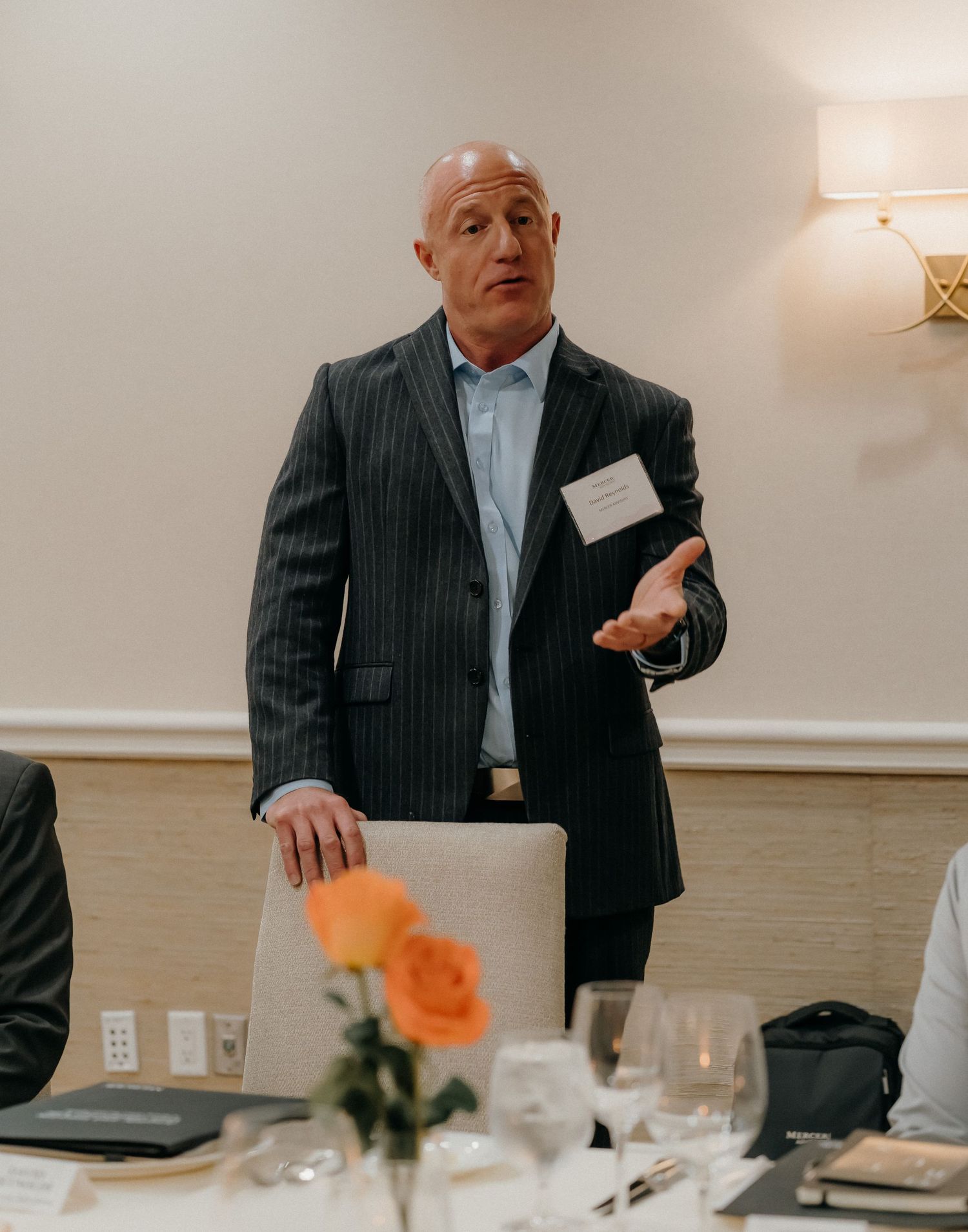 A person in a pinstripe suit gestures while standing at a formal dining table with orange flowers and place settings.