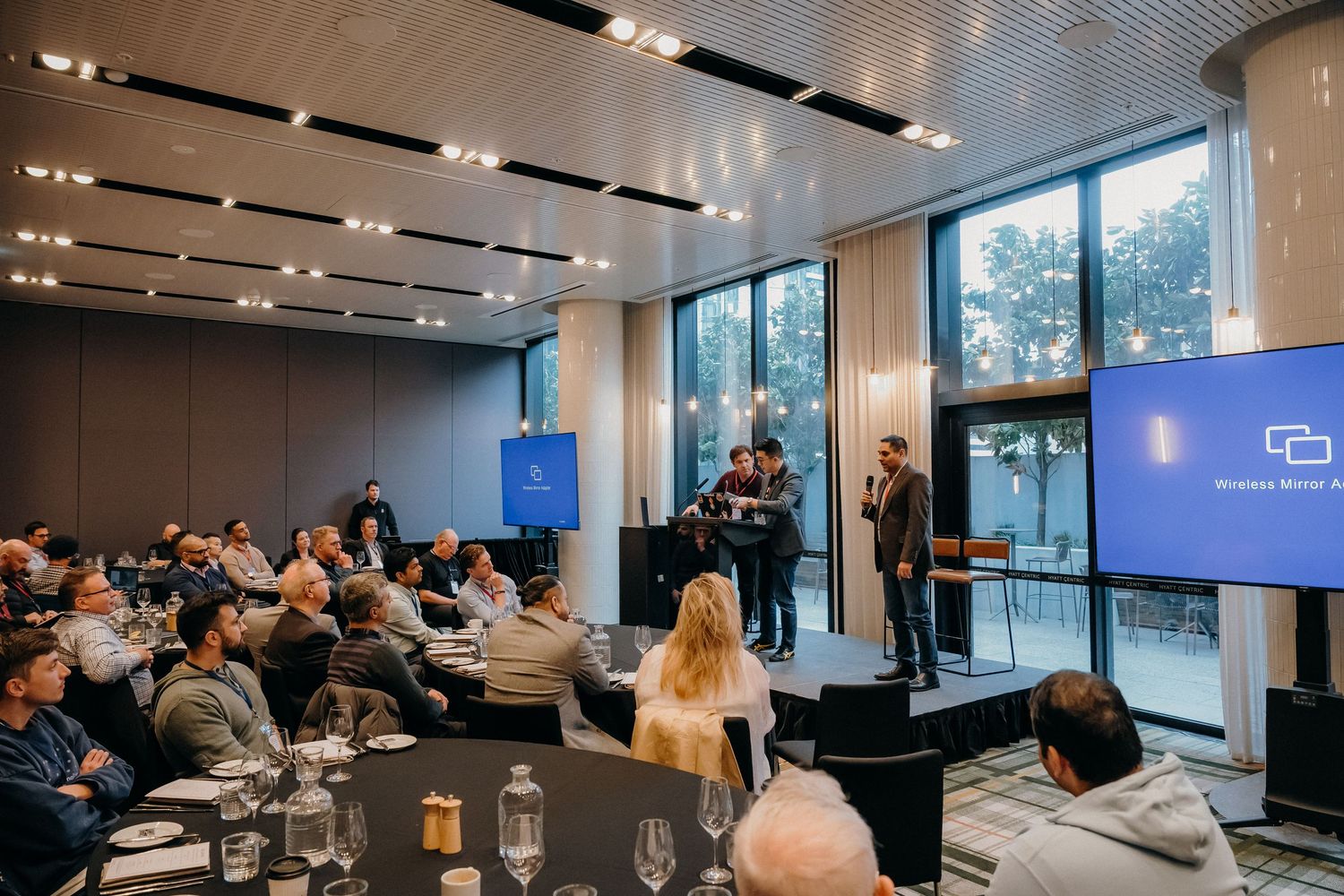 Business conference attendees gather in a modern meeting room with floor-to-ceiling windows and presenters on stage.