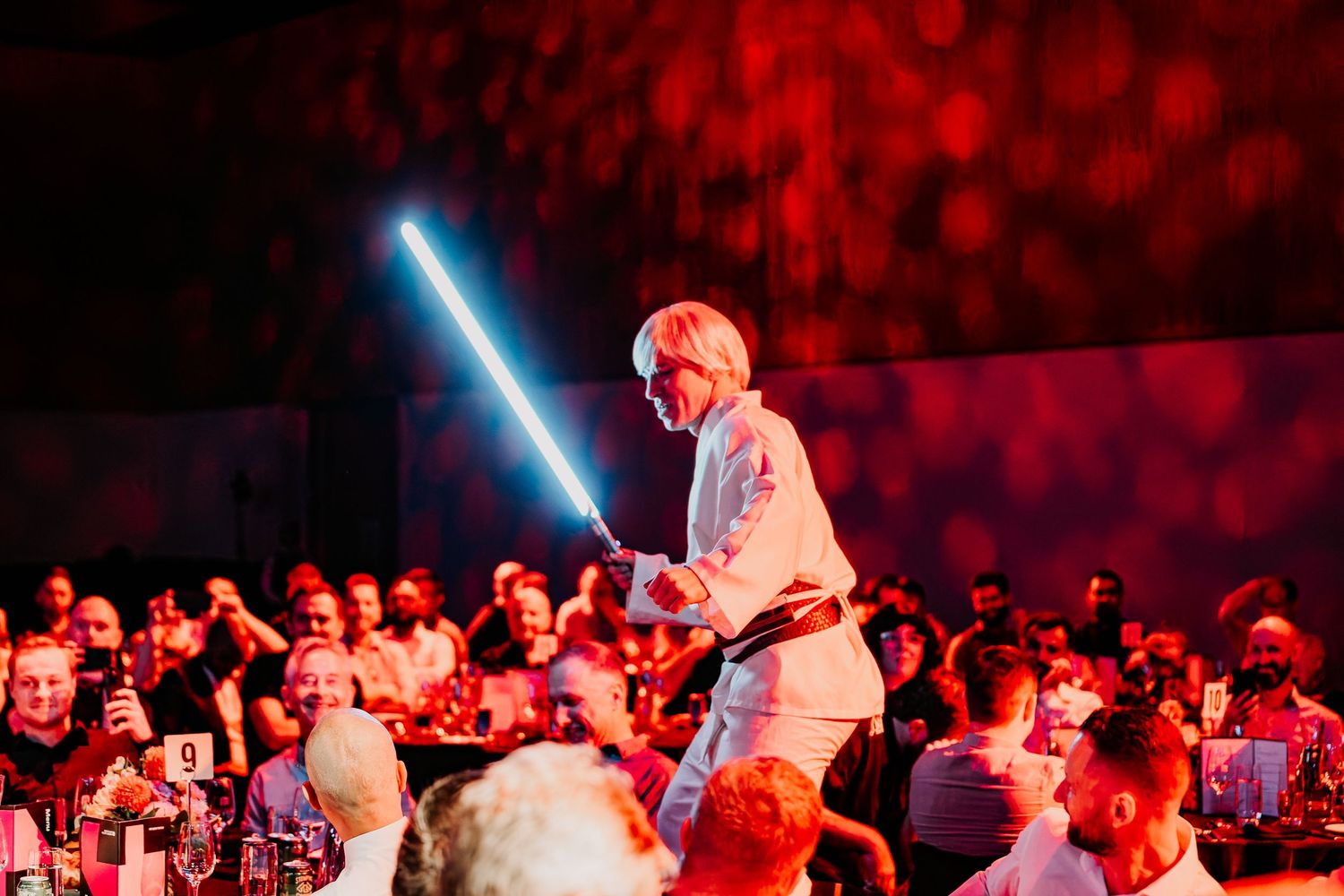 Concert performer in white shirt plays guitar on stage with dramatic red lighting and audience in view.