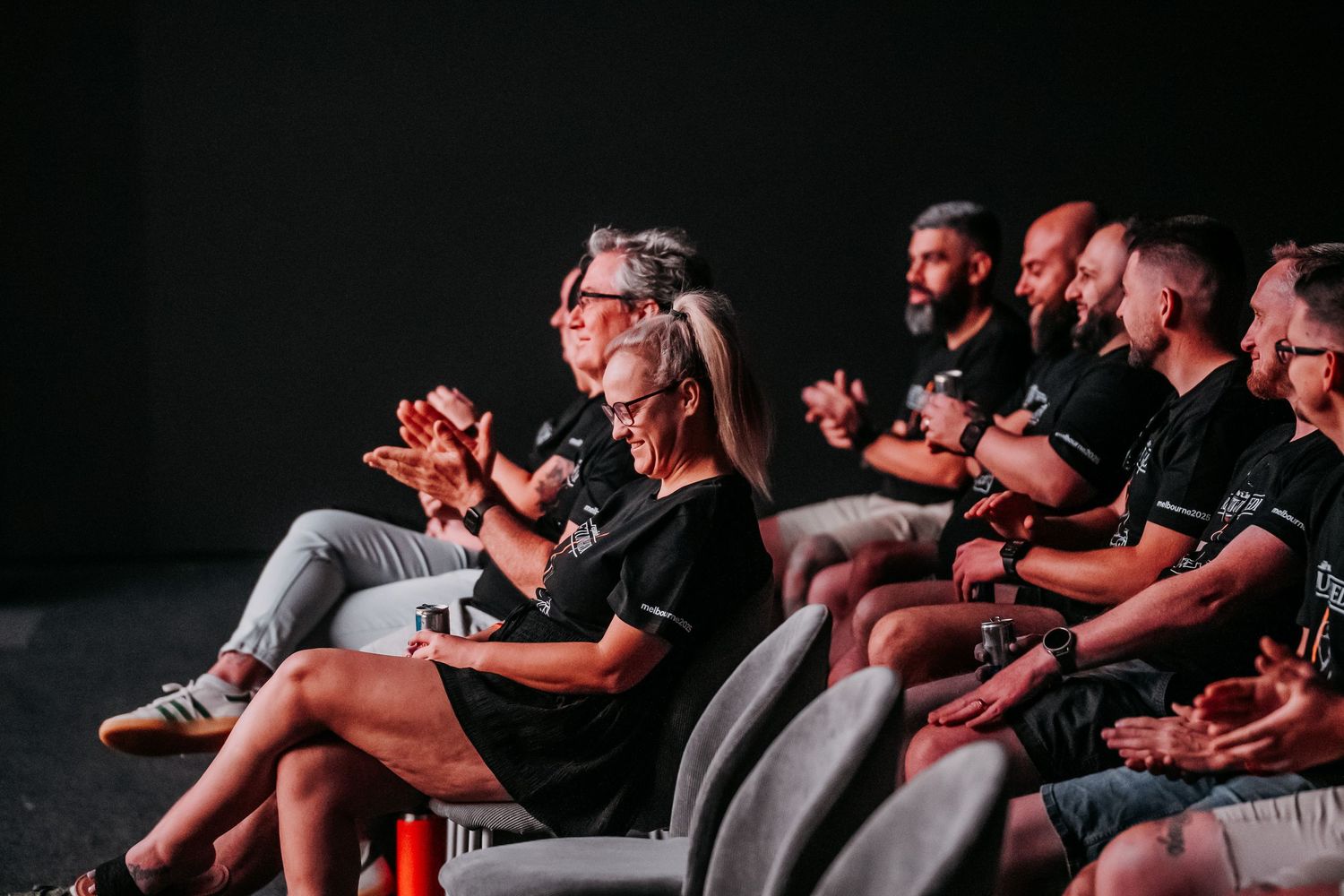 Audience members in dark attire sit in theater seats applauding during a performance.