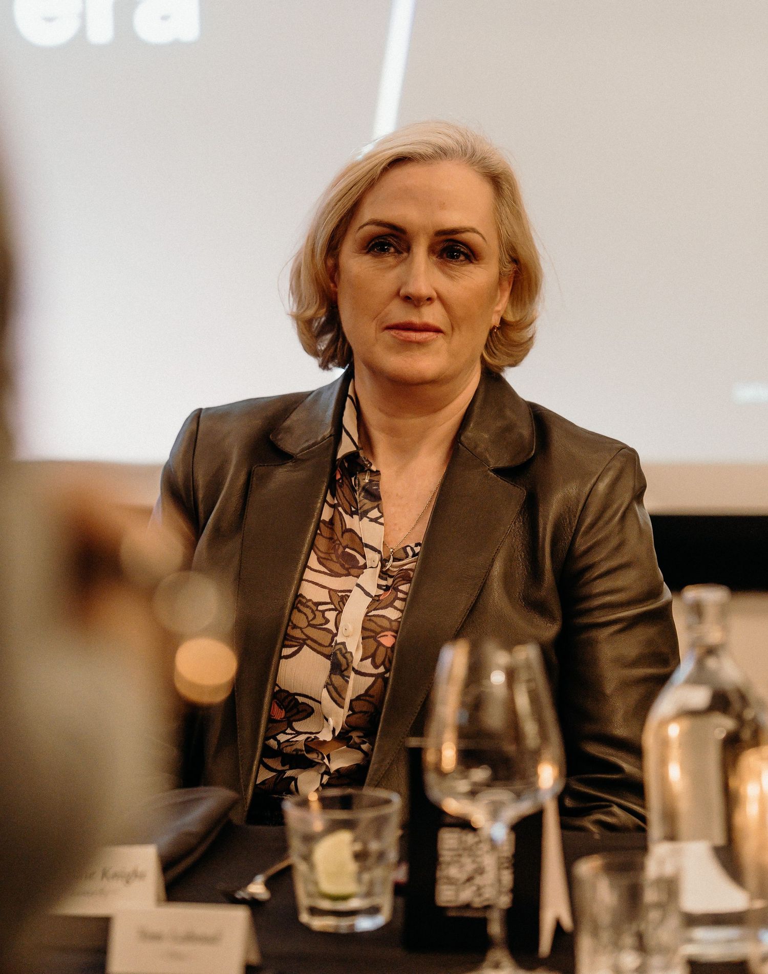 Professional wearing a dark blazer and patterned blouse sits at a formal business meeting with wine glasses on the table.