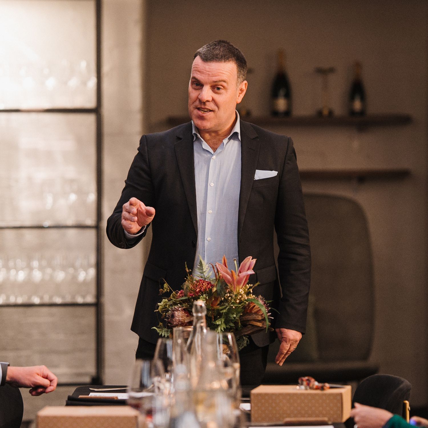 Professional speaker in dark suit gestures while giving presentation at formal dining event.