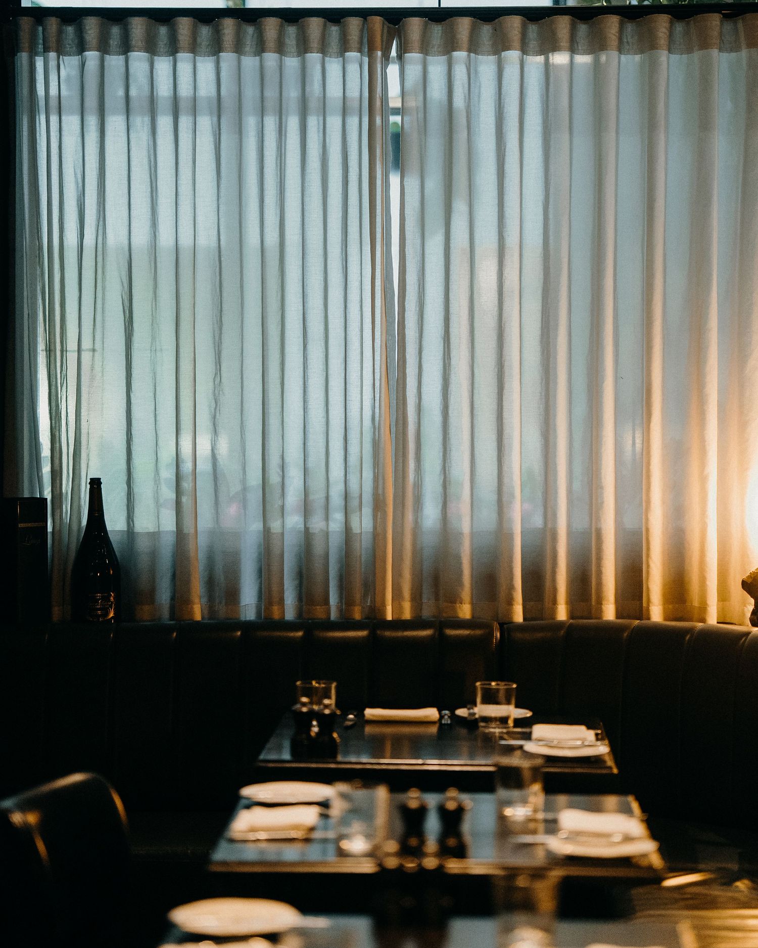 Moody restaurant interior with translucent curtains backlit by warm lighting and an elegant table setting in the foreground.