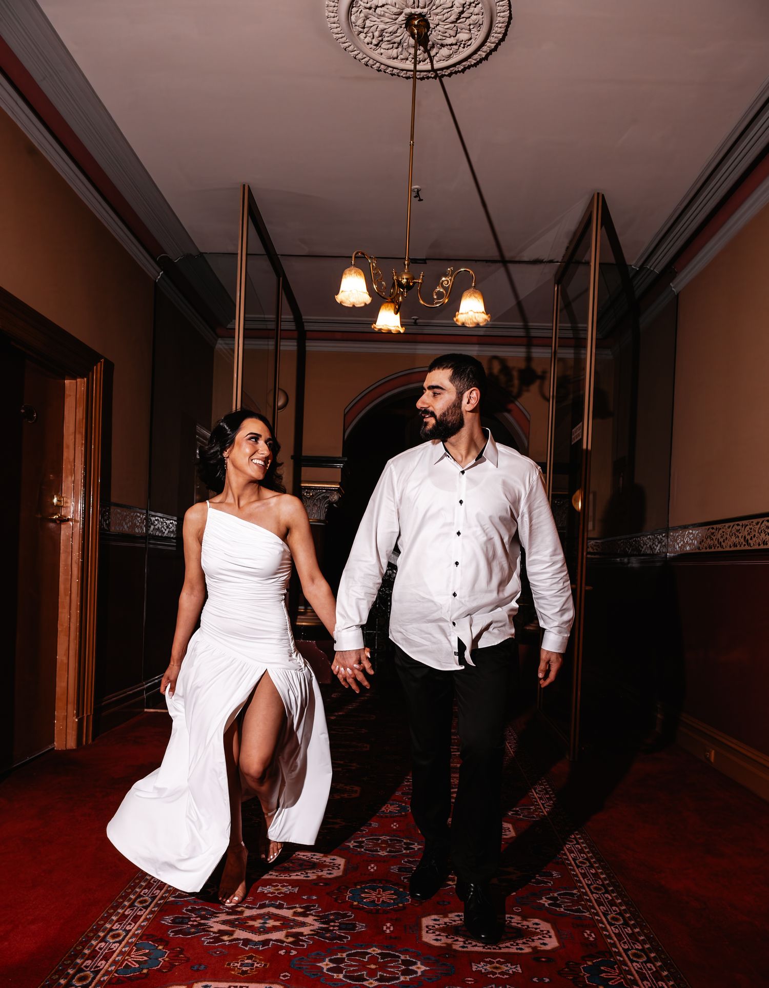 A couple in white and black formal attire dances gracefully on a red carpet in a dimly lit hallway.