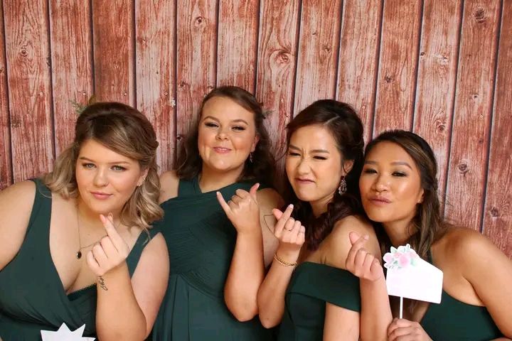 Four friends pose together in front of a rustic wooden wall wearing dark formal dresses.