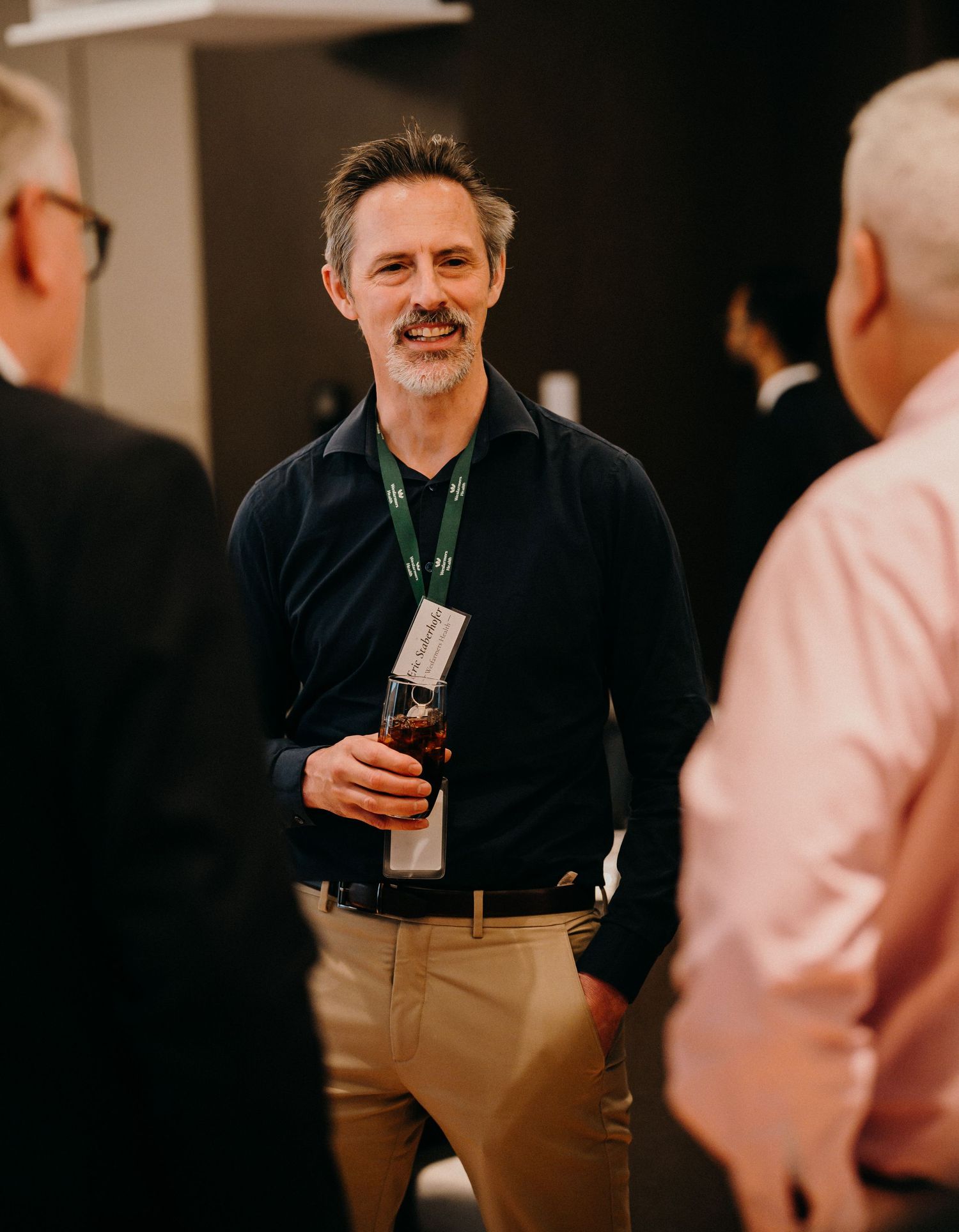 Professional in a dark sweater and khakis holds a beverage while engaged in conversation at a networking event.