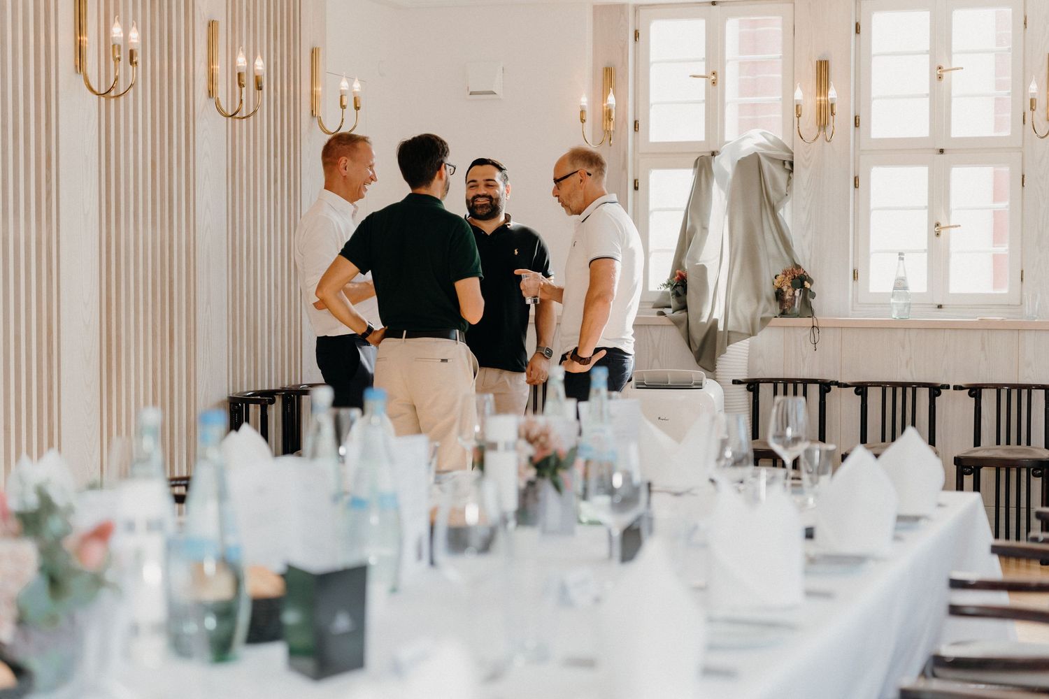 Elegantly set banquet tables in a bright venue with three men in smart casual attire conversing in the background.