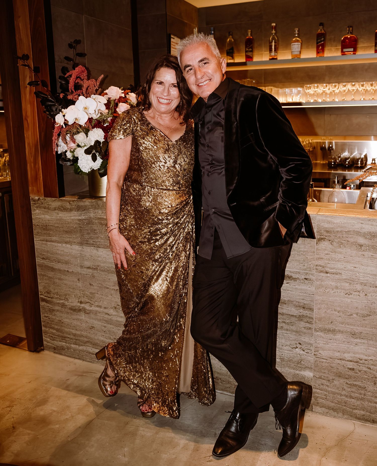 A couple poses at an elegant bar, with the woman wearing a gold sequined gown and the man in a black velvet jacket.