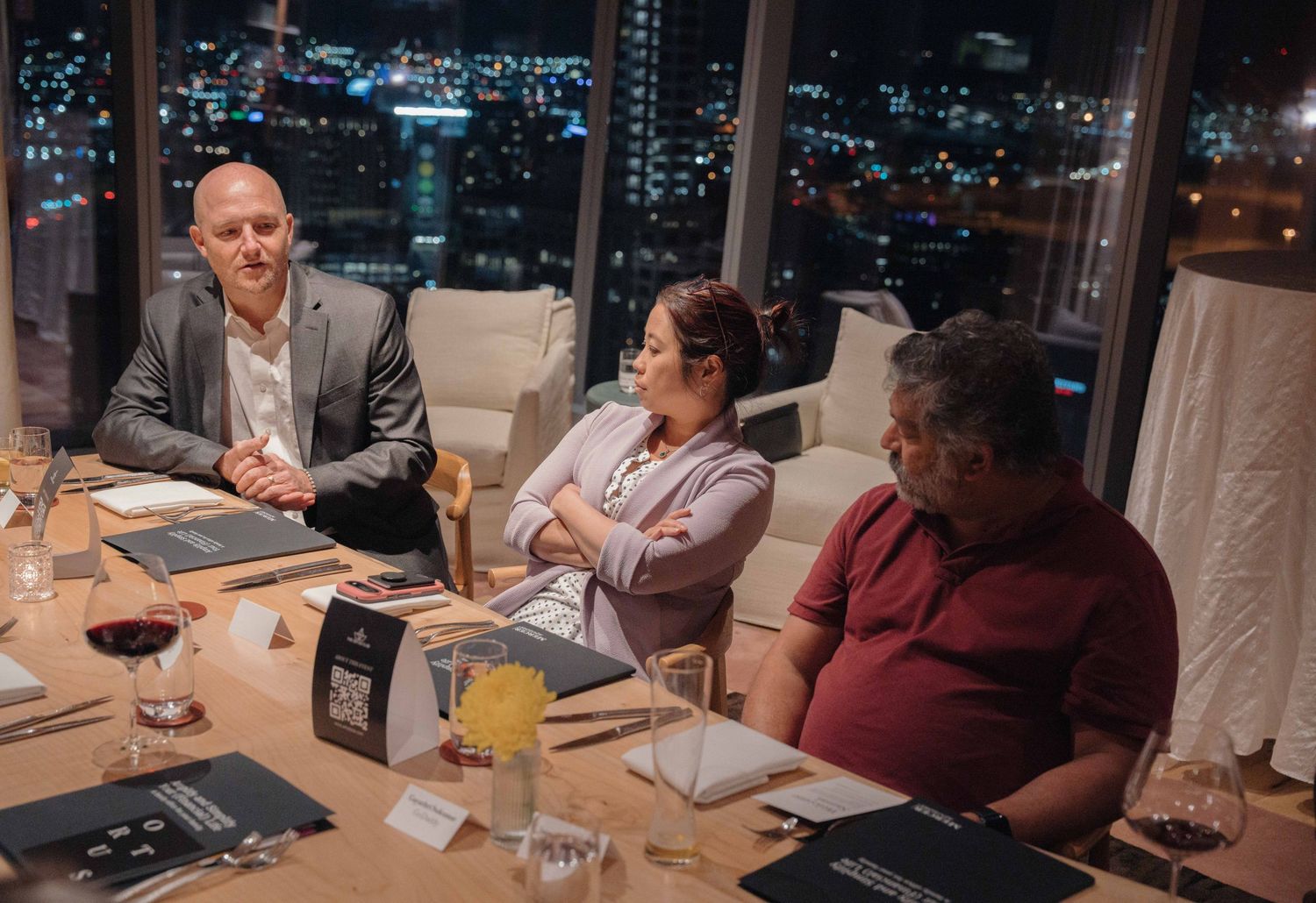 Three people having a business meeting at night in a high-rise restaurant with city lights visible through the windows.