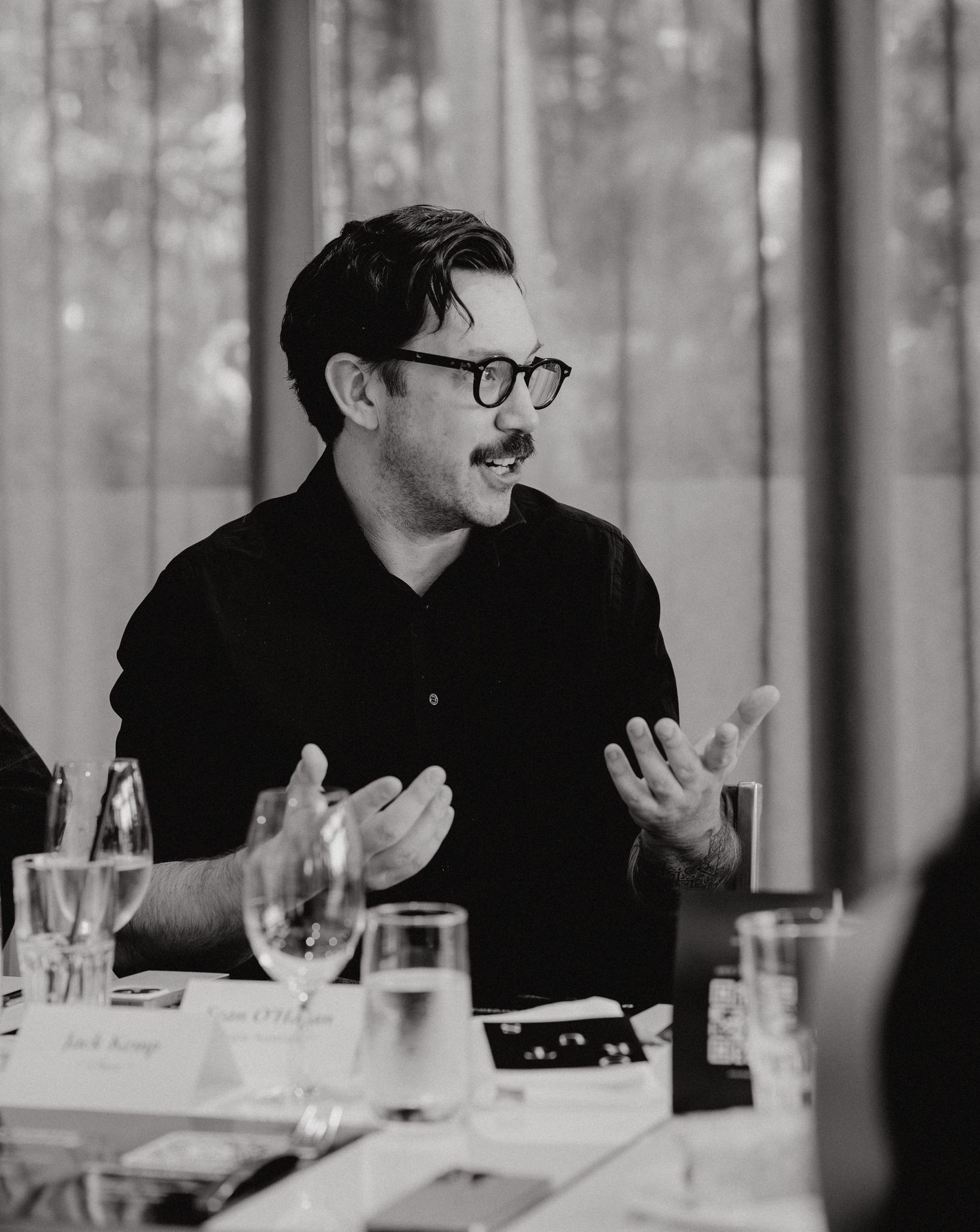 Black and white photo of someone in glasses laughing expressively while seated at a dining table with glassware.