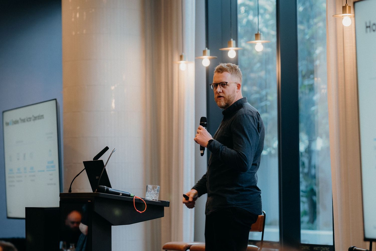 A person in a dark suit gestures while giving a business presentation in a modern office meeting room.