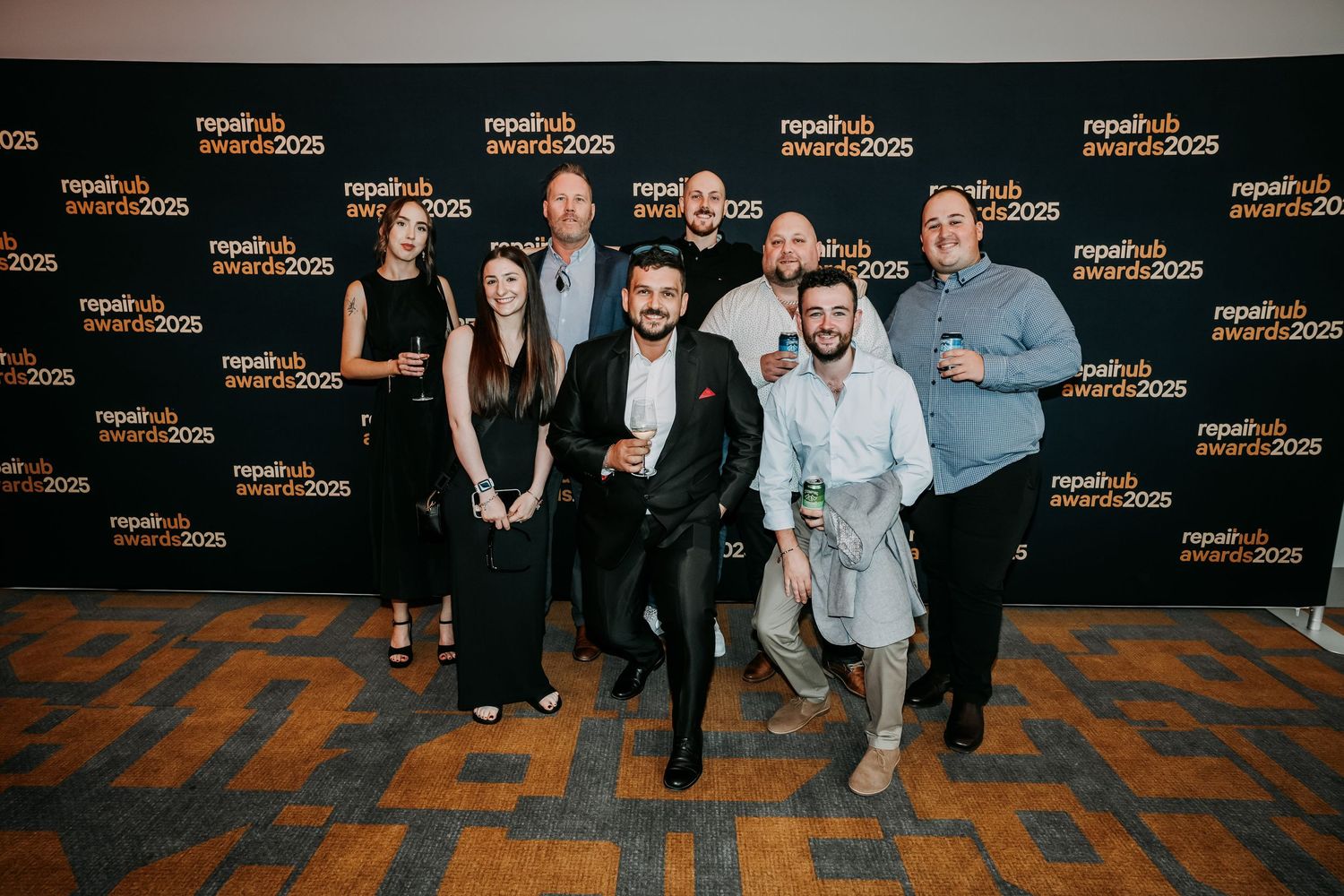 A group of people pose together in formal attire against a branded backdrop with orange and black geometric patterns.