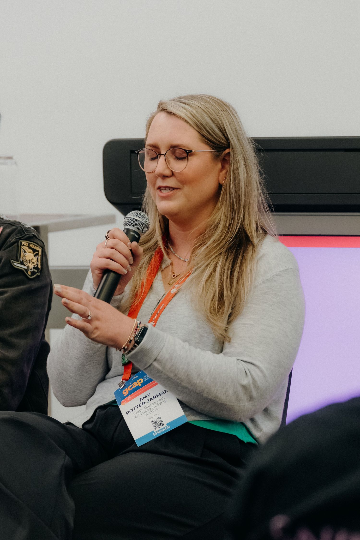 A speaker wearing glasses and a gray sweater presents at a conference while holding a microphone.