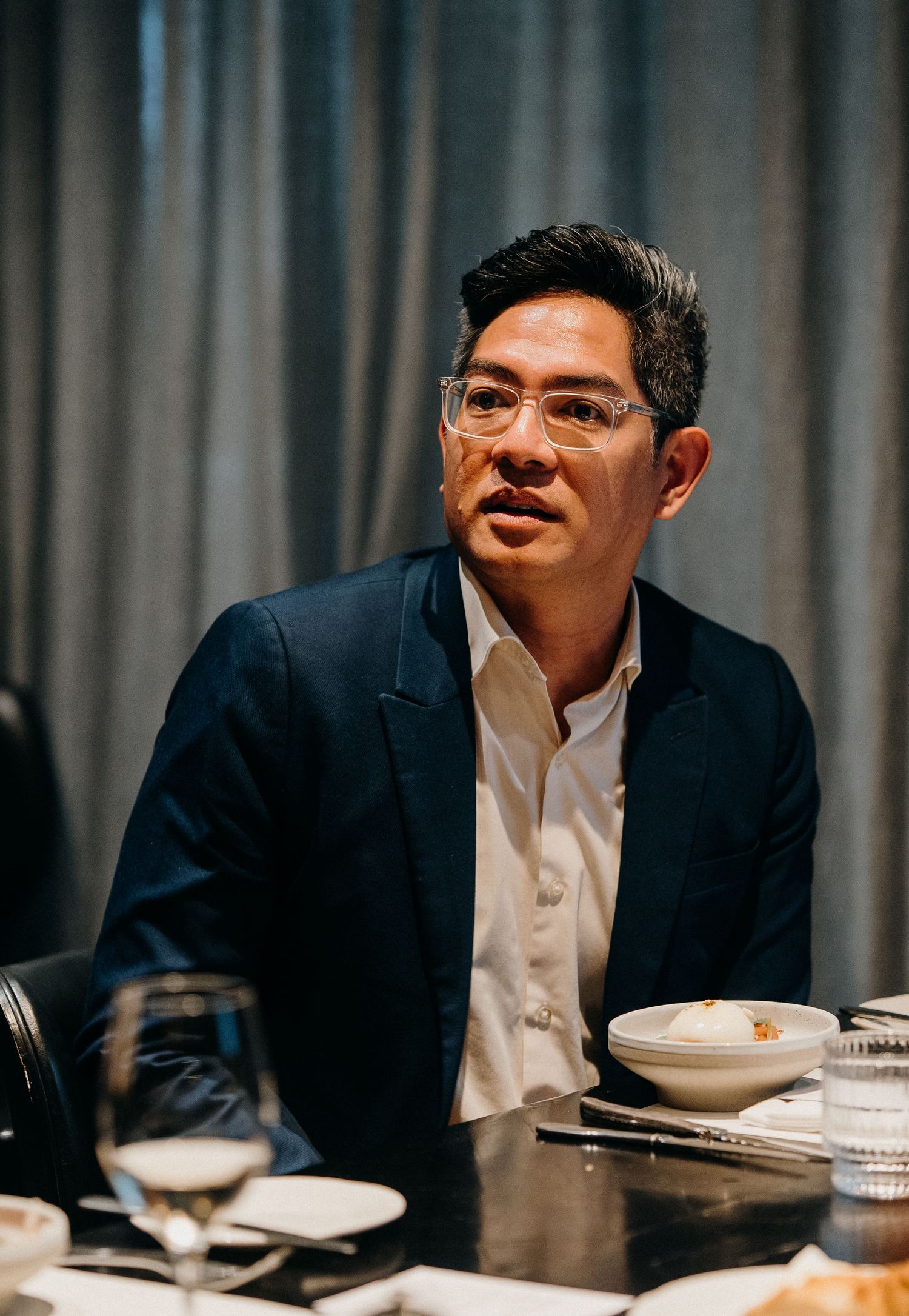 Professional in a navy suit and glasses sits at a dining table with white plates and cups in a dimly lit setting.