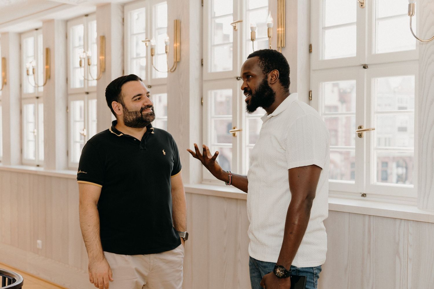 Two men having a conversation in a bright, modern room with white paneled walls and elegant wall sconces.
