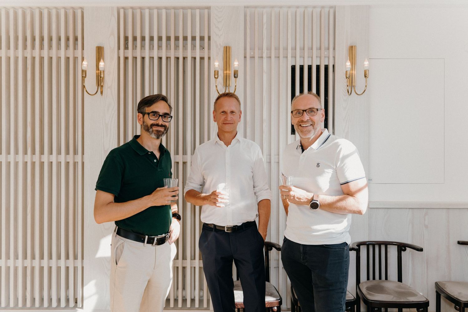 Three smiling men in smart casual attire holding drinks in an elegant white interior with gold wall sconces.