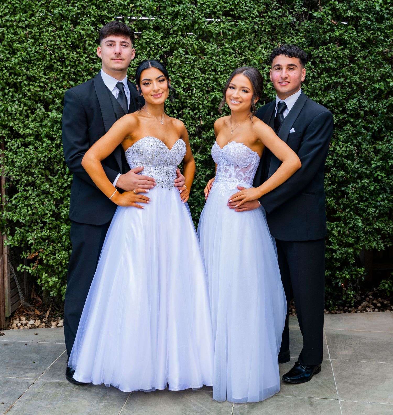 Wedding party in formal attire poses against lush green hedge background at outdoor venue.