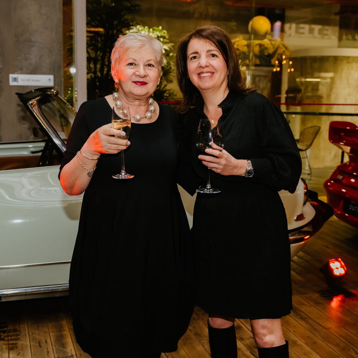 Two women in black dresses smile while holding wine glasses at an evening event with a classic car in background.