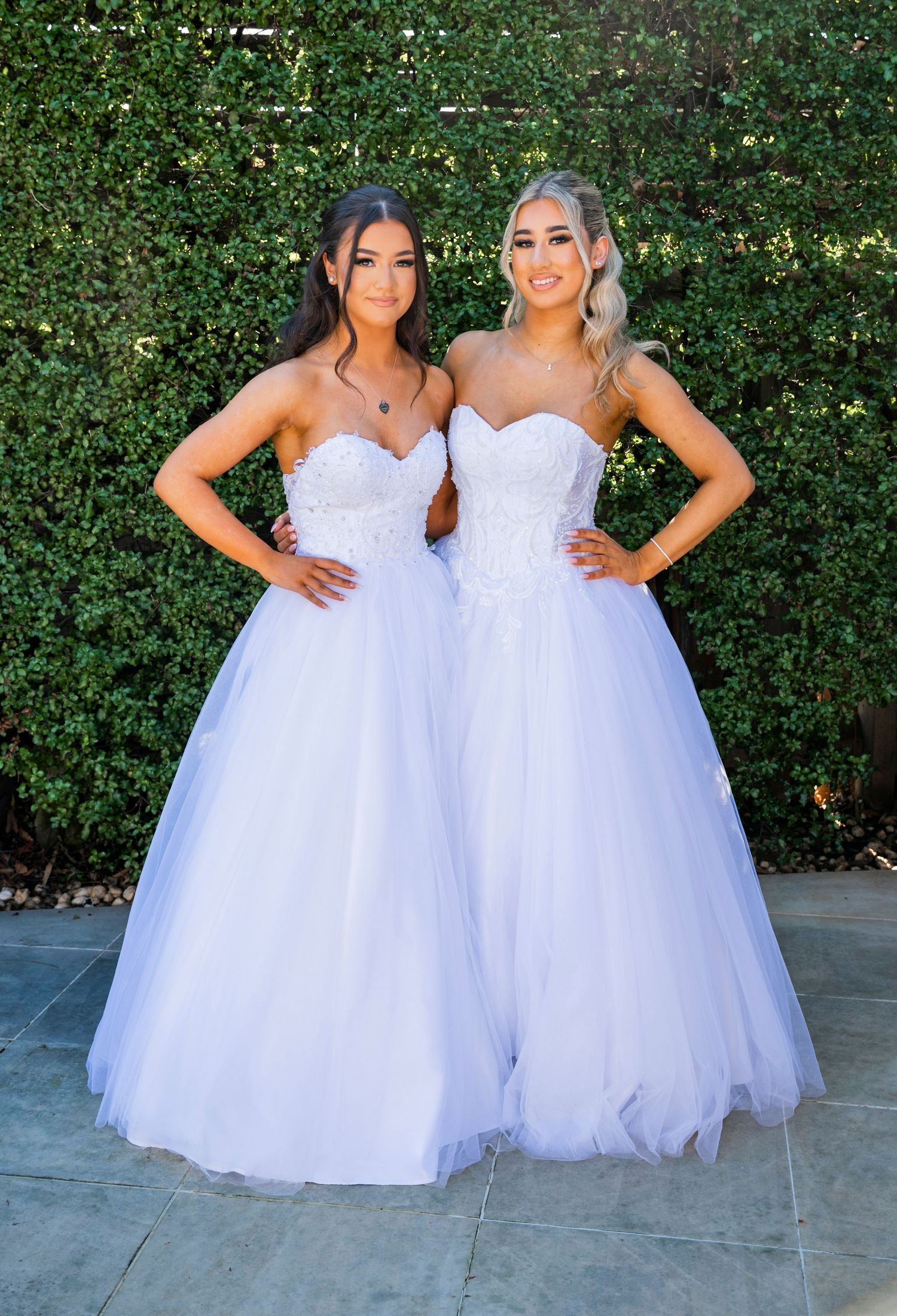 Two people in matching white sweetheart neckline ball gowns pose together against a hedge background.