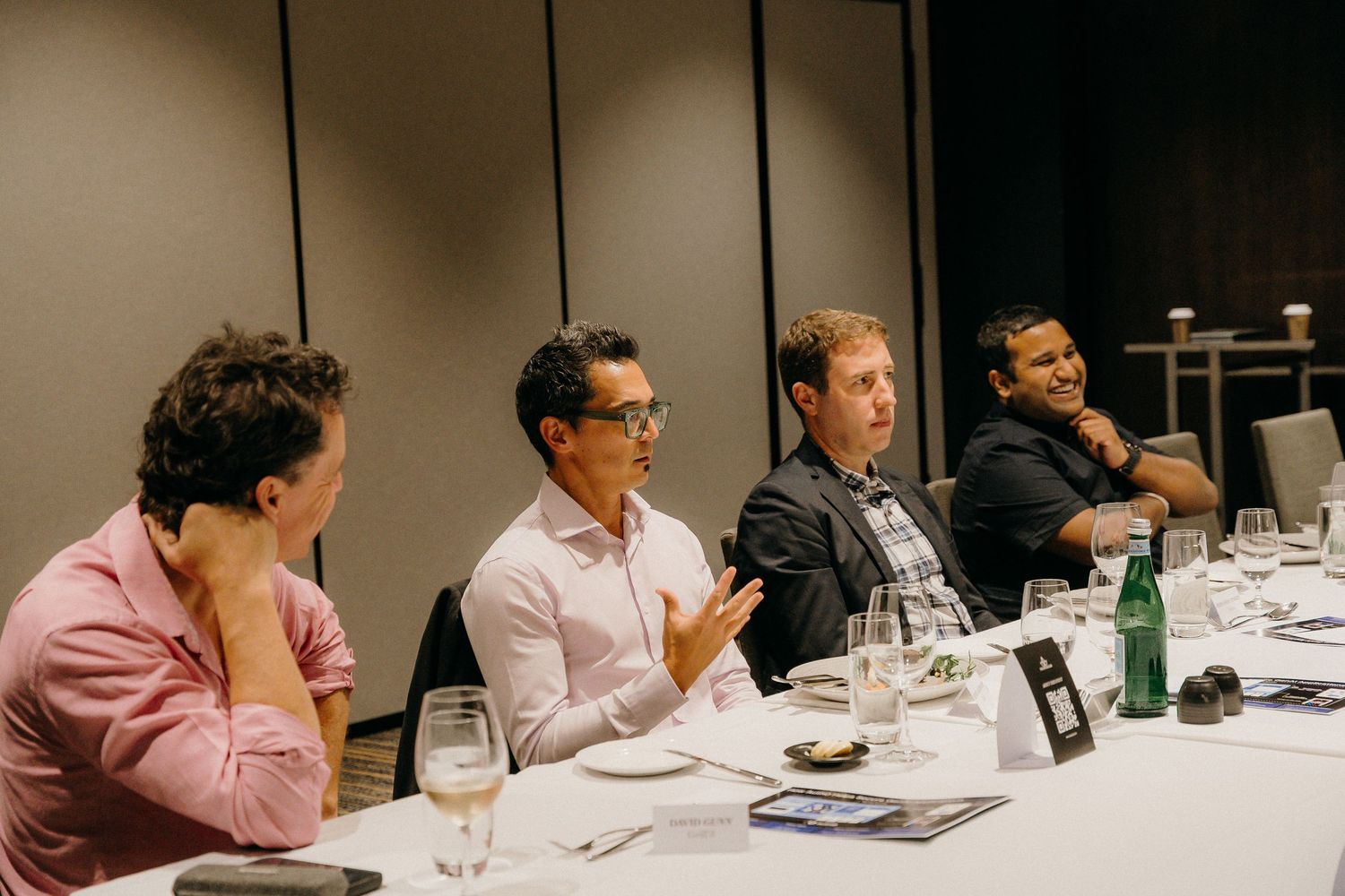 Four businessmen engaged in discussion at a corporate dinner meeting around a white tablecloth table.