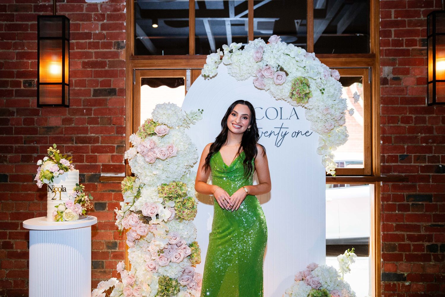 Person in emerald green evening gown poses near floral wall decorations at an elegant brick venue.