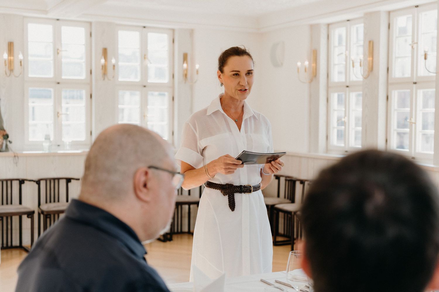 Woman in white dress presenting to audience in bright elegant room with large windows.