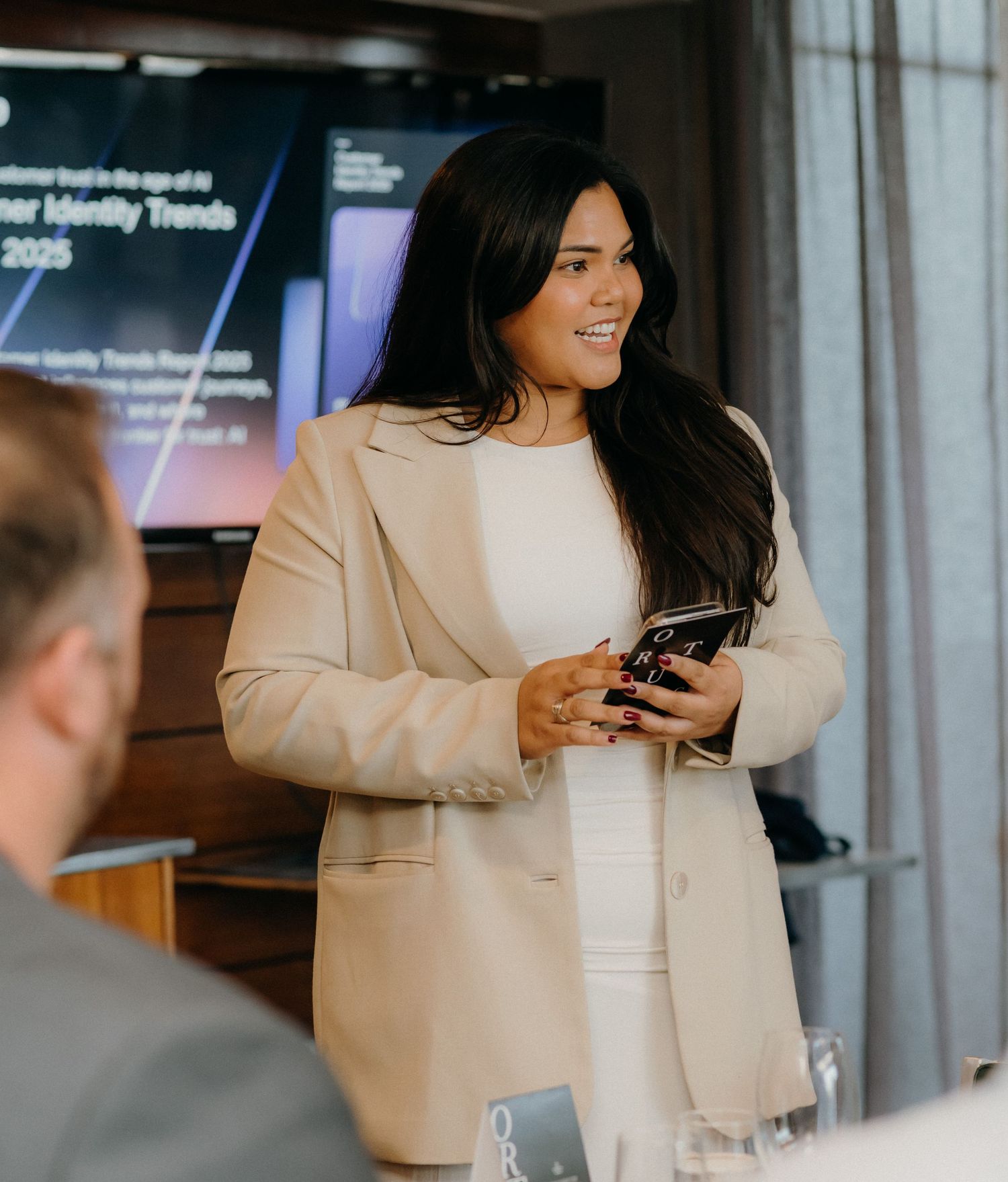 Professional in a beige blazer checks mobile phone while standing in modern office setting.