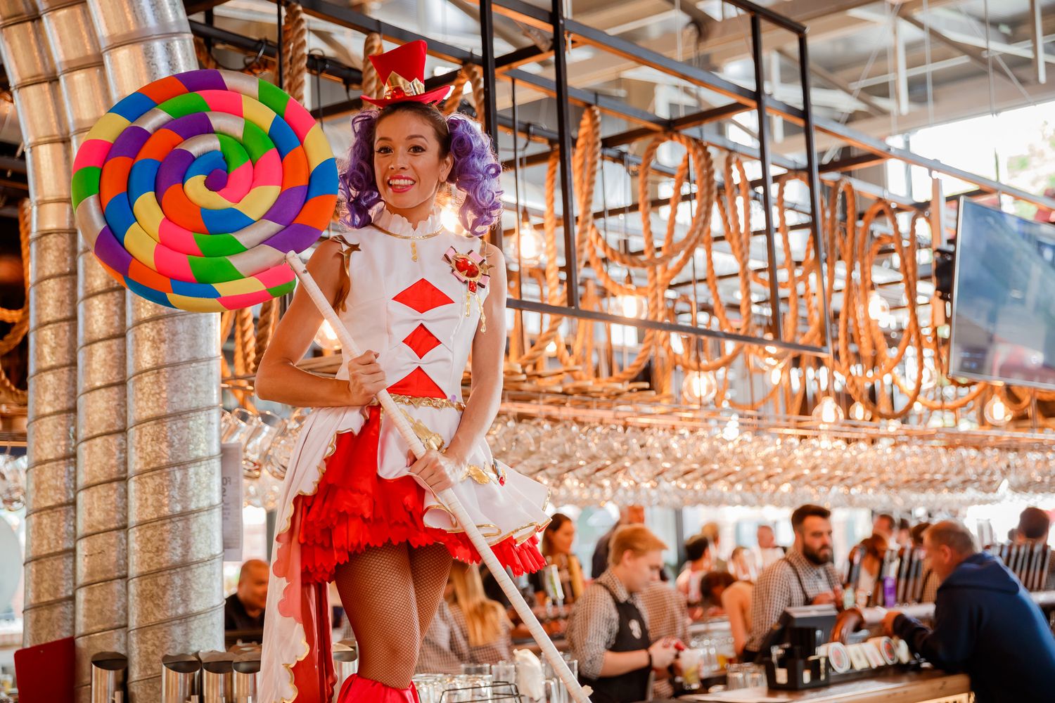 Performers in colorful costumes dance and entertain guests at an indoor venue with wooden beams and string lights.