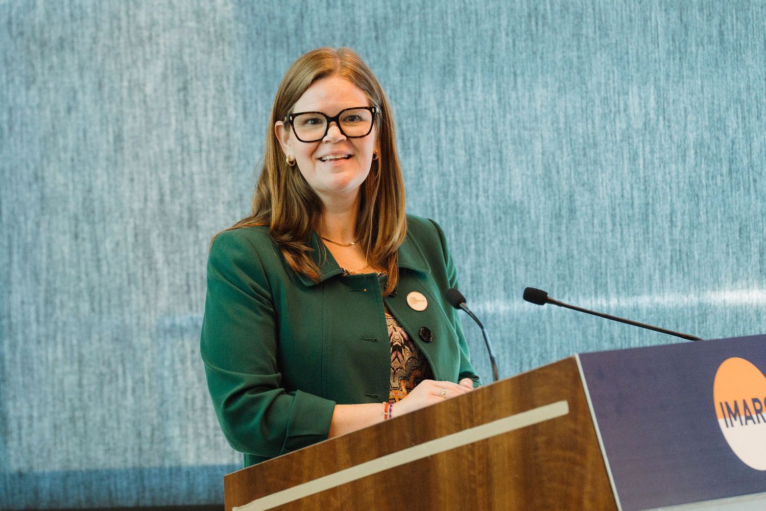 Speaker stands at podium wearing glasses and green sweater while addressing audience at professional event.