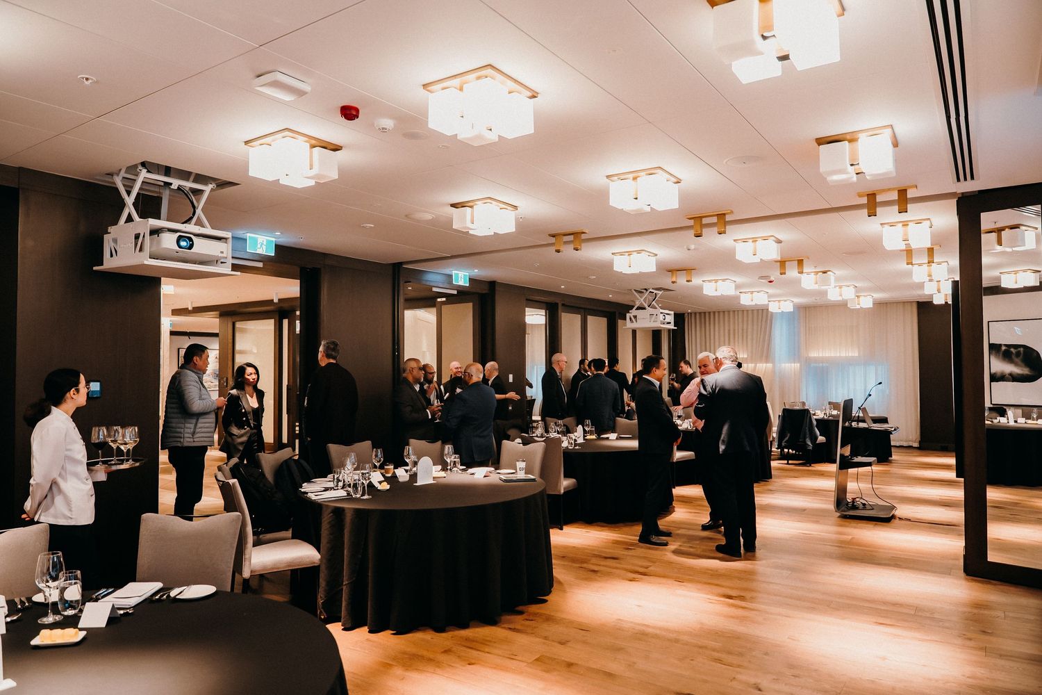 People gather in an elegant event space with dark walls, hardwood floors and modern square ceiling lights.